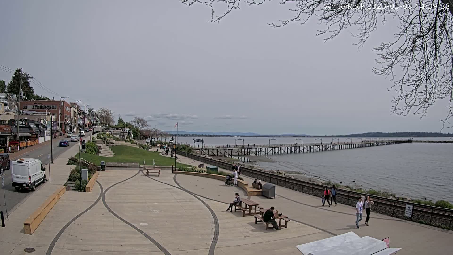 A long pier extends into a calm, gray-toned body of water under an overcast sky, with people strolling along a paved waterfront path lined with benches and buildings.