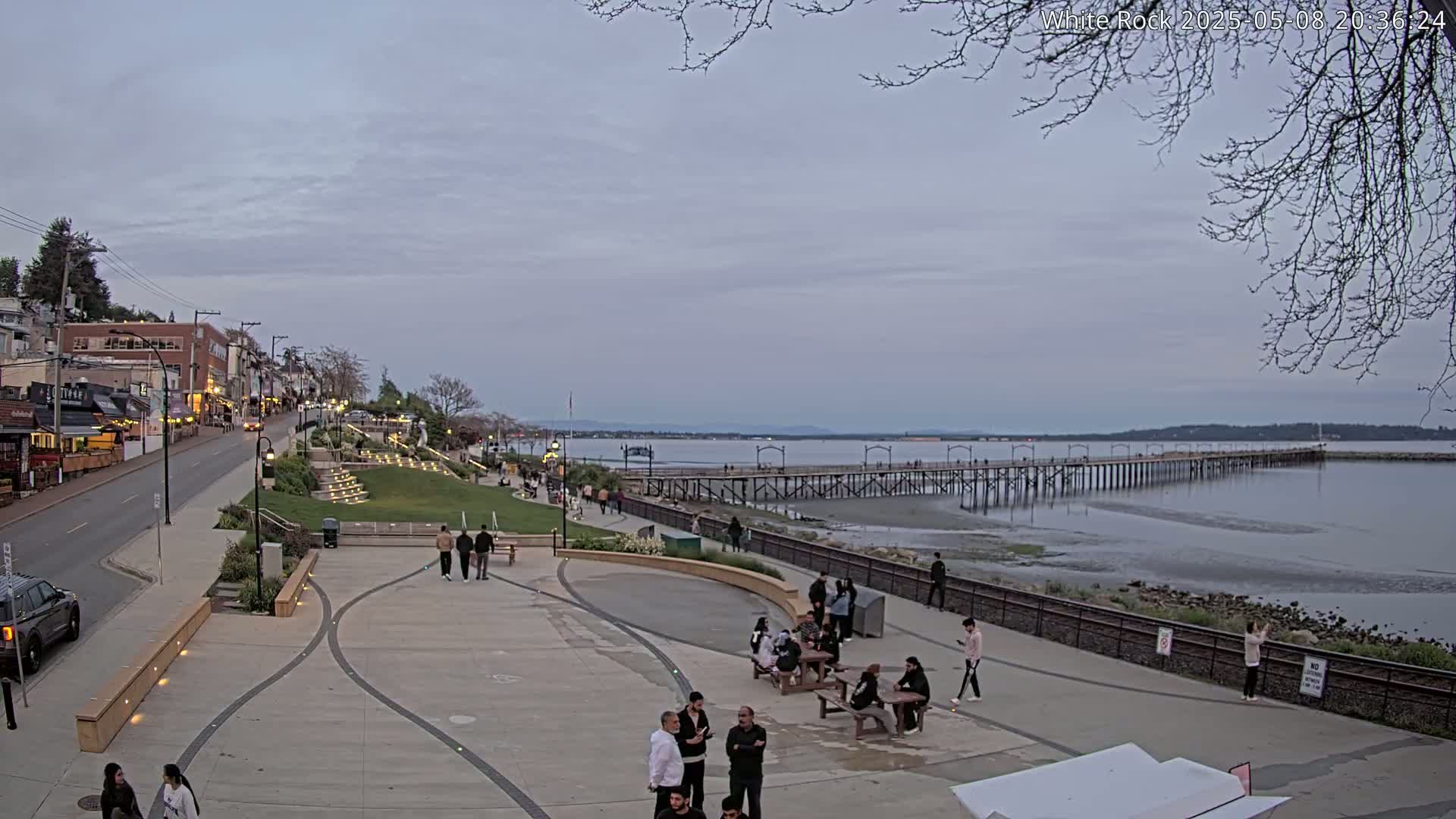 A waterfront town square with a long pier extending into a calm, overcast bay, featuring people strolling and sitting at picnic tables.