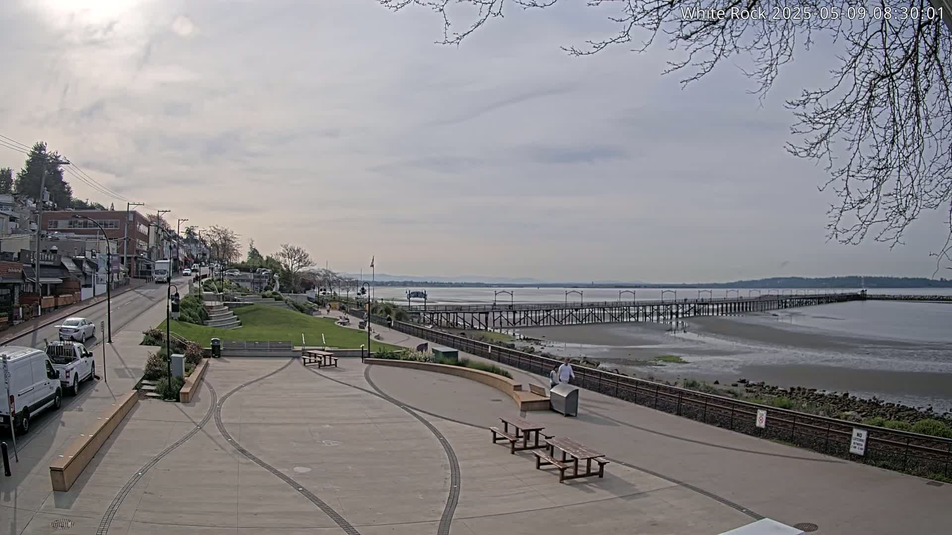 A paved waterfront plaza with picnic tables overlooks a long pier extending into a calm, gray-sky day's ocean.