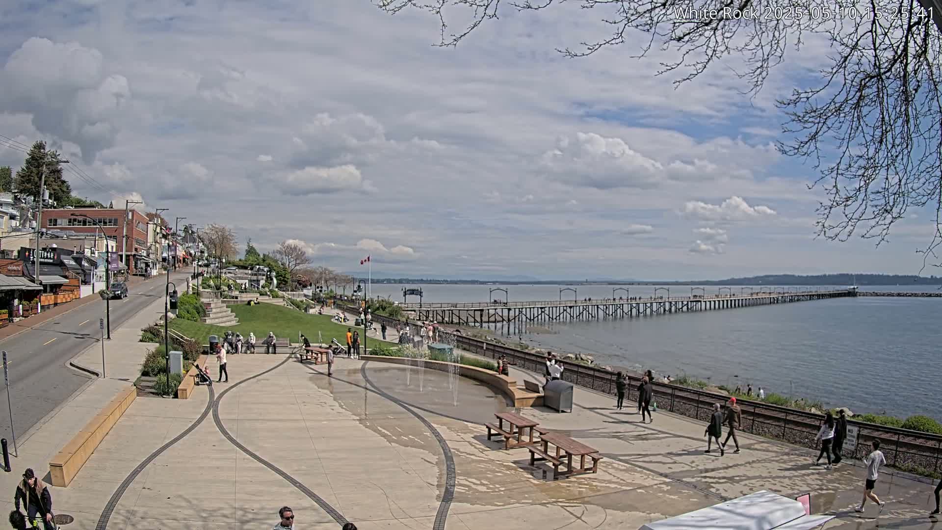 A partly cloudy day reveals a waterfront scene with a long pier extending into a calm body of water, a paved promenade lined with people, and a town with buildings in the background.