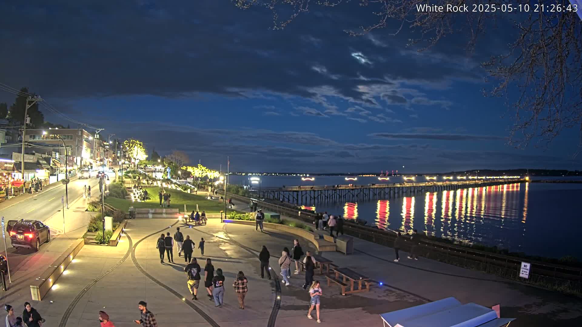 Under a partly cloudy night sky, people stroll along a waterfront promenade near a brightly lit pier extending into a calm, dark-blue body of water, with buildings and streetlights illuminating the scene.