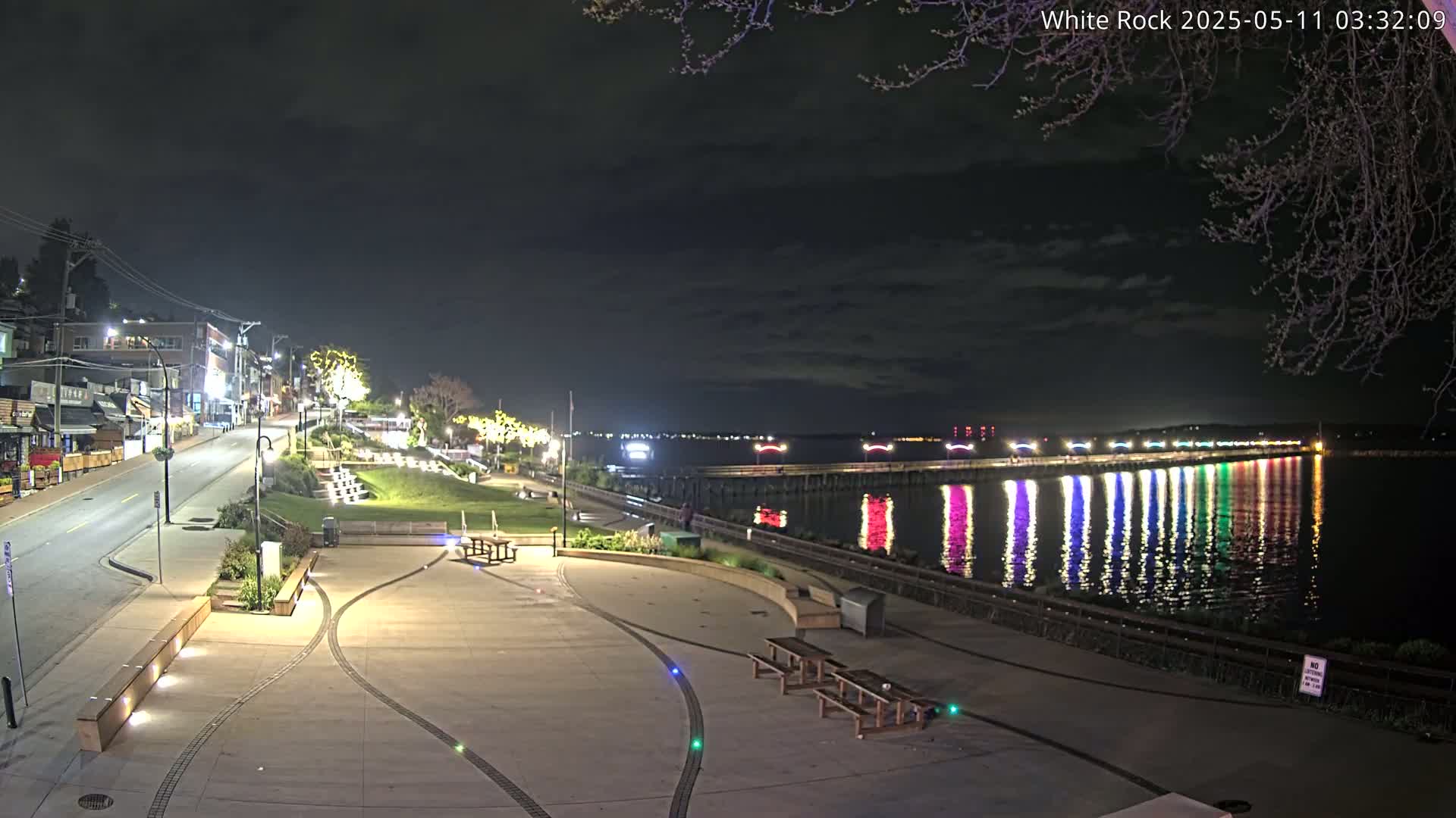 A nighttime view of a waterfront plaza with benches and lights, a street leading to buildings, and a pier with colorful lights reflected in calm water under a partly cloudy sky.