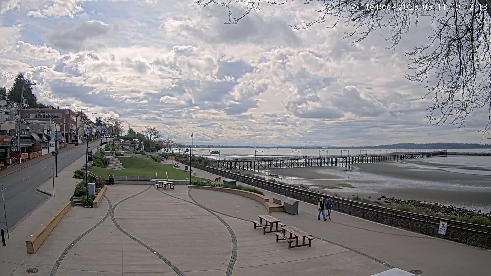 A paved waterfront plaza with picnic tables overlooks a long pier extending into a partly exposed bay under a partly cloudy sky.