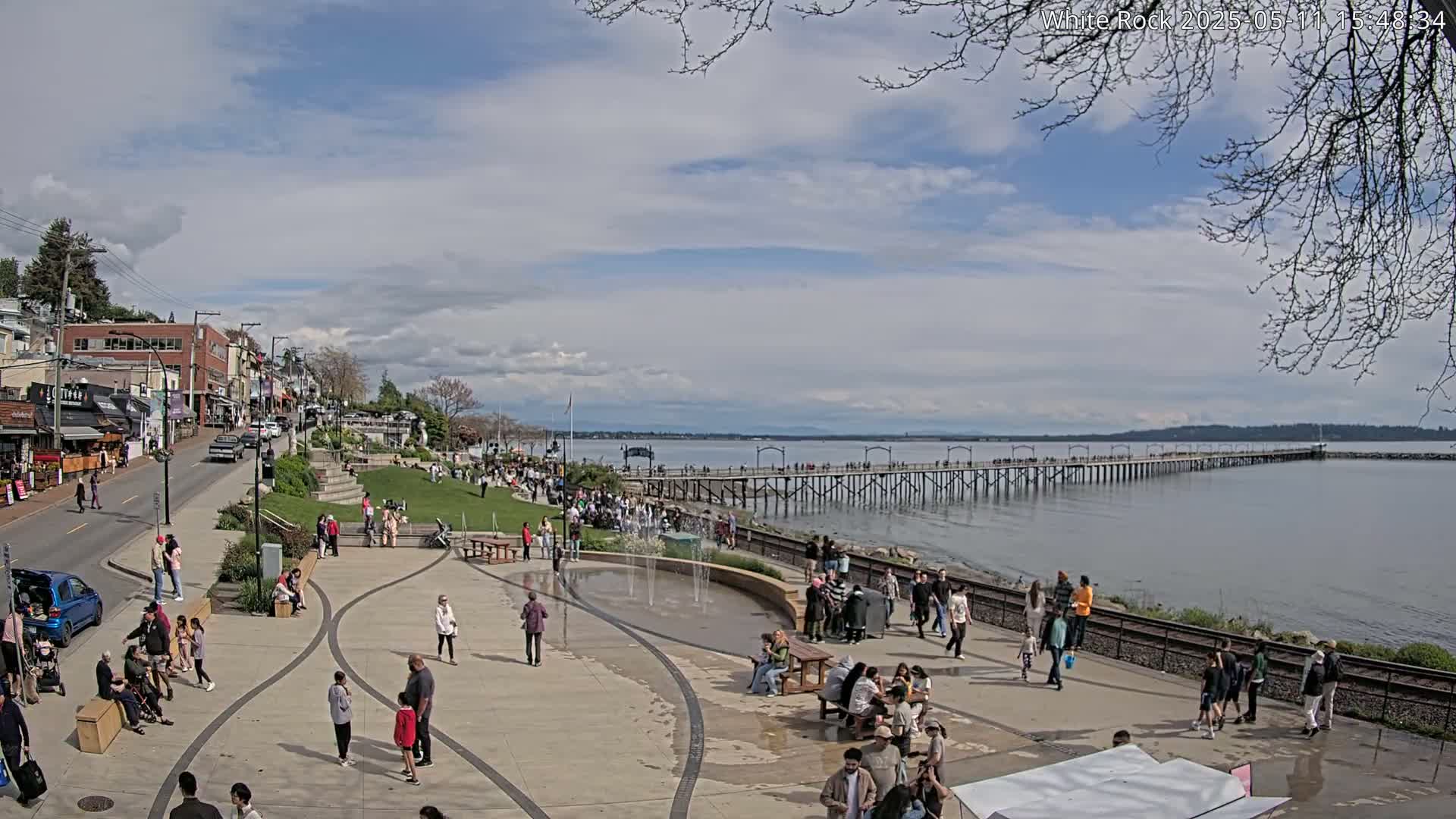 A partly cloudy day reveals a waterfront scene with a long pier extending into the calm water, where many people stroll along a paved plaza with small fountains, near shops and buildings.
