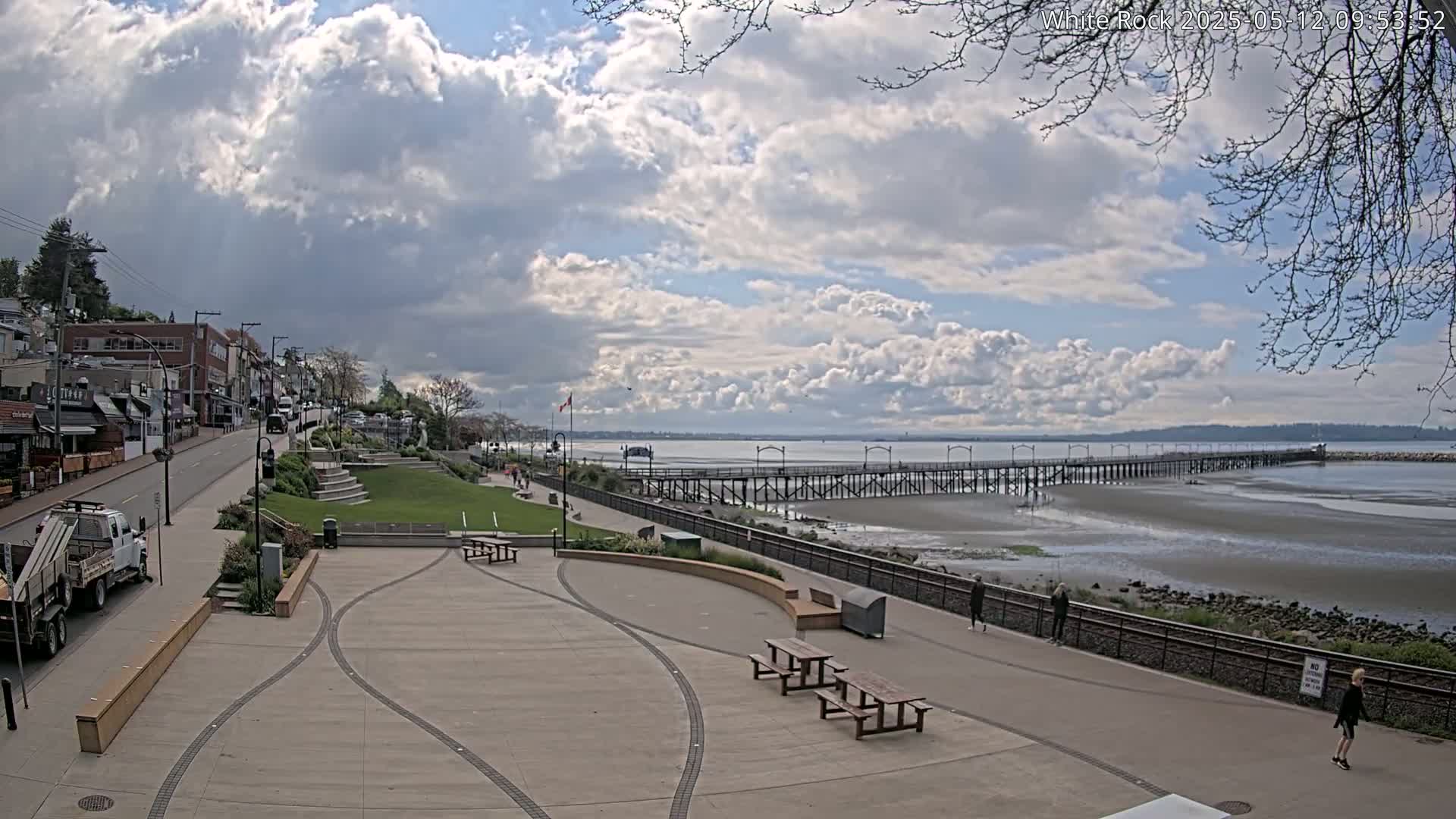 A partly cloudy day shows a waterfront scene with a long pier extending into a calm bay, a paved walkway, and a plaza with picnic tables.