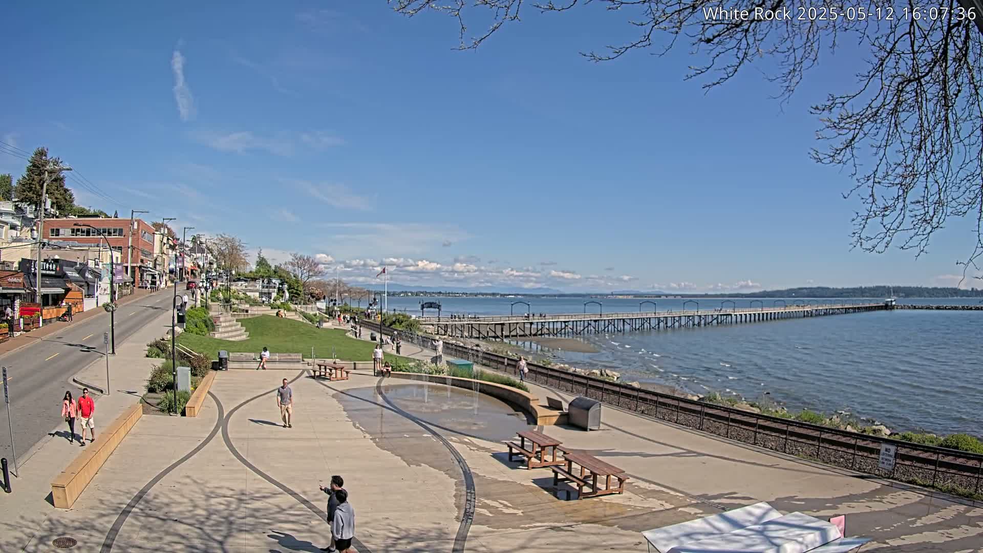 A waterfront town scene on a sunny day shows a long pier extending into a calm bay, with people strolling along a paved walkway near shops and a small park.