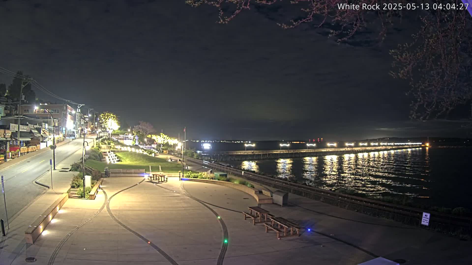 A nighttime view of a waterfront plaza with a long pier extending into a calm body of water under a dark sky.