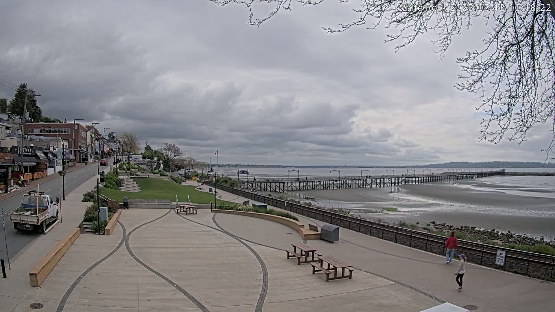 A paved waterfront plaza with picnic tables overlooks a long pier extending into a calm, grey-cloudy day sky over a wide, exposed sandy beach.