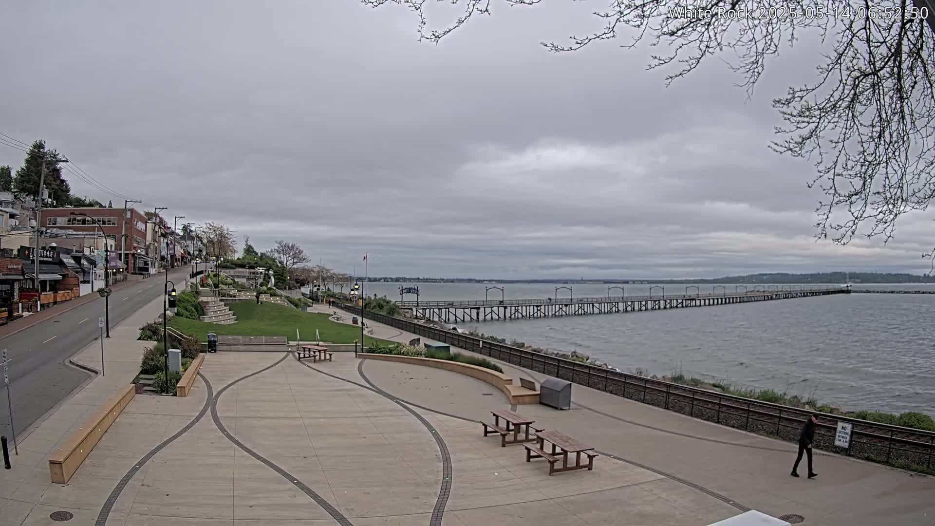 A long pier extends into a calm, grey-sky ocean next to a paved waterfront area with picnic tables and a street lined with shops.