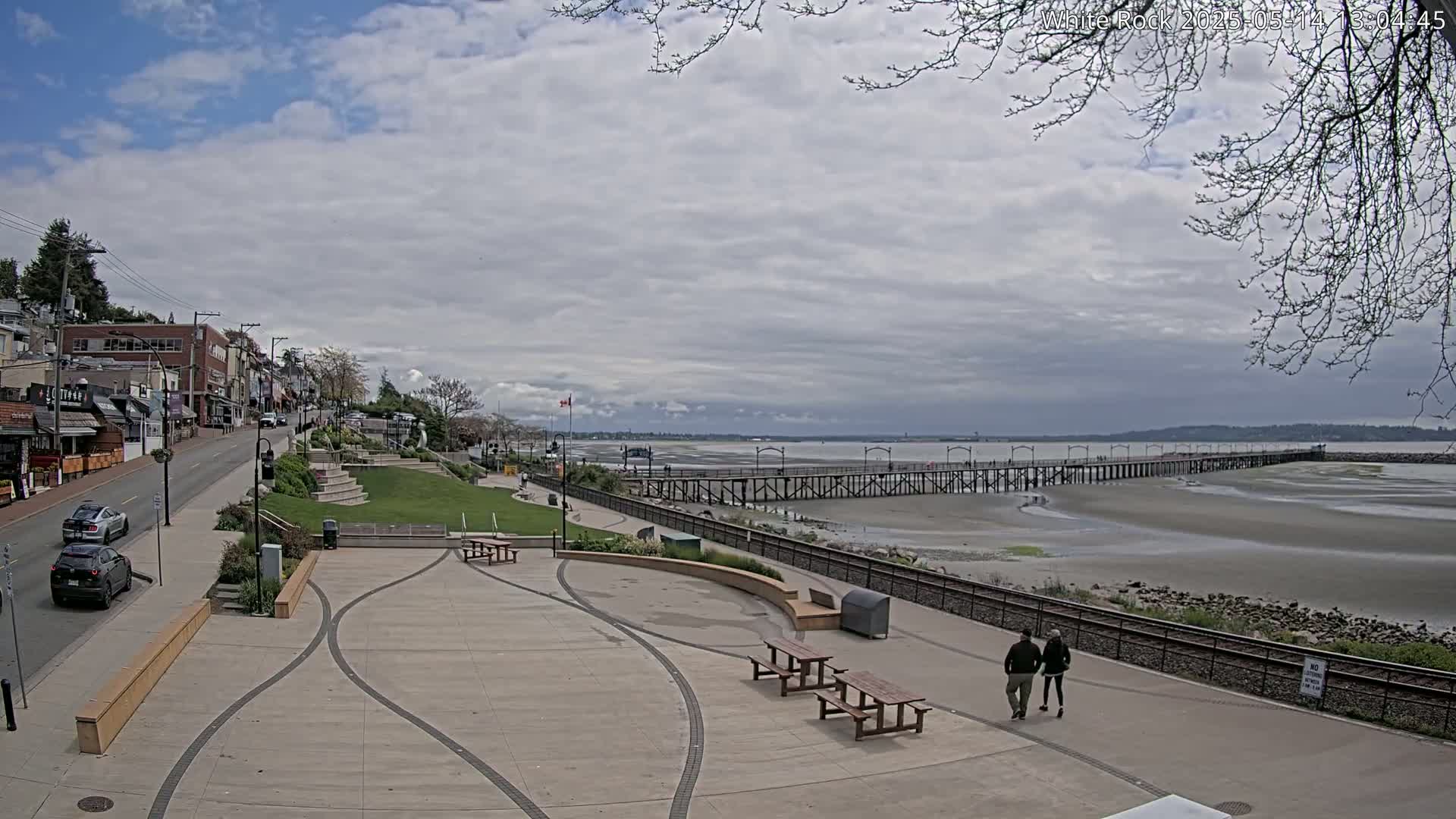 A paved waterfront plaza with picnic tables overlooks a long pier extending into a wide, exposed sandy beach under a partly cloudy sky.