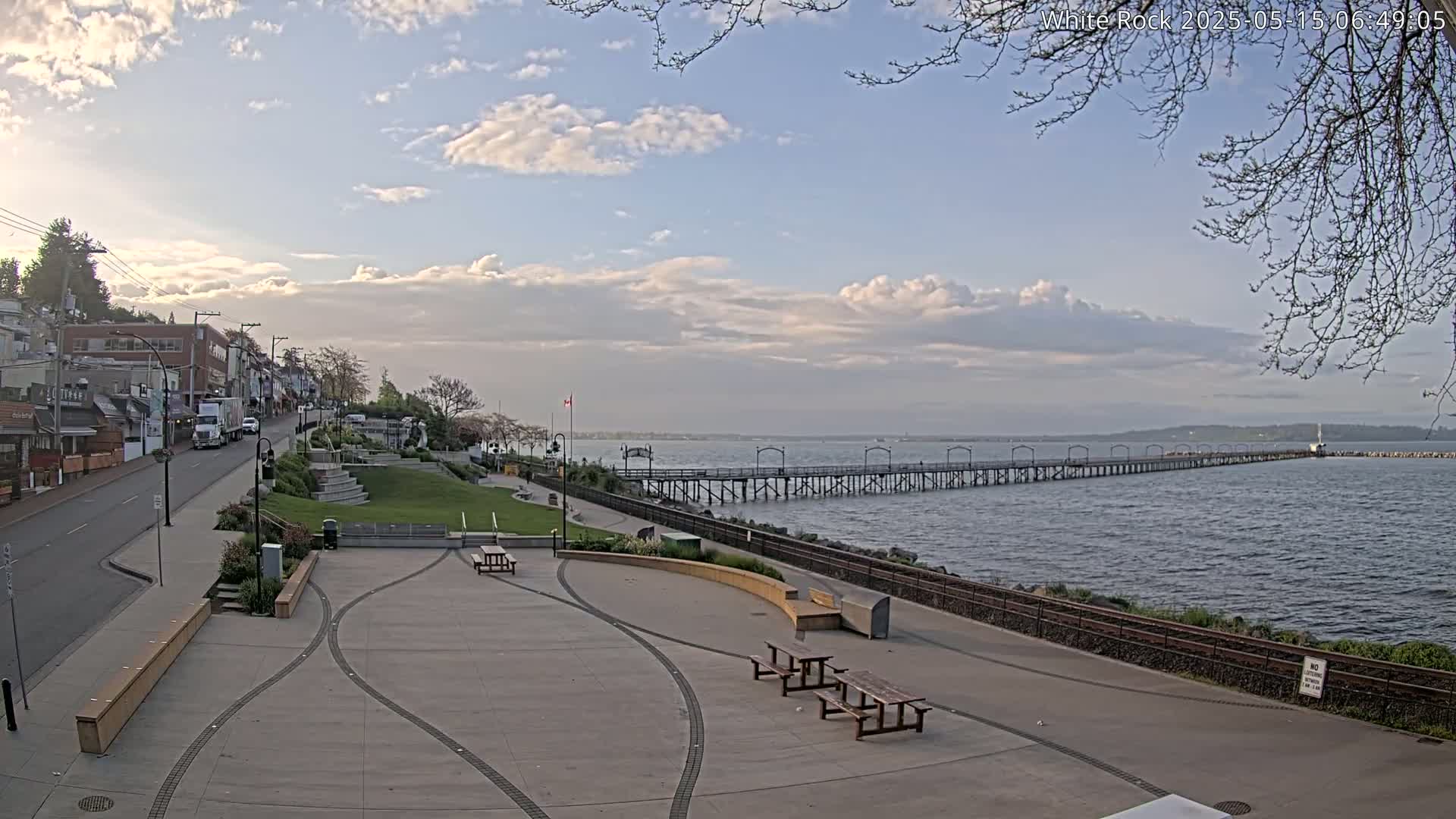 A partly cloudy day reveals a seaside town with a long pier extending into calm water, a paved waterfront area with picnic tables, and buildings lining a street uphill.