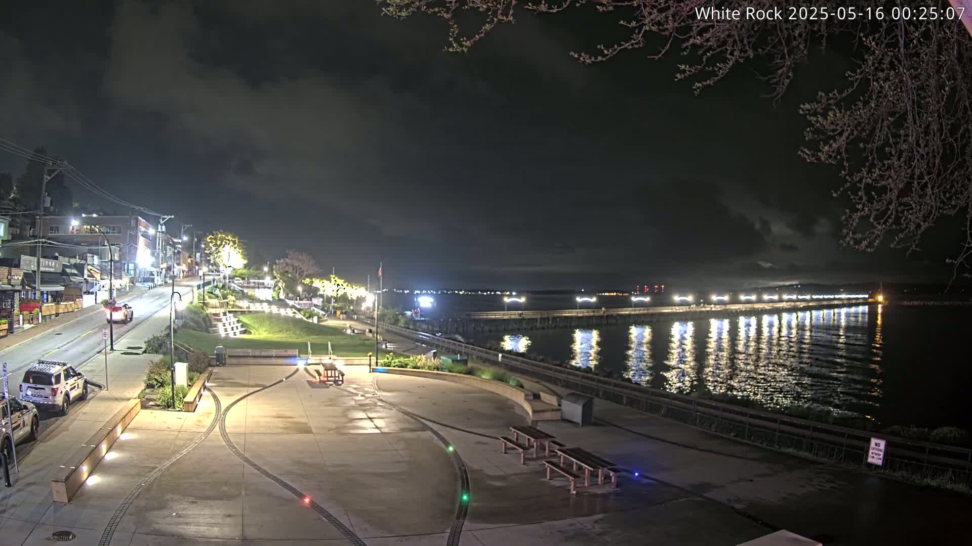 A nighttime view of a waterfront area shows a paved plaza with benches, a street with vehicles, and a long pier extending into calm water under a partly cloudy sky.