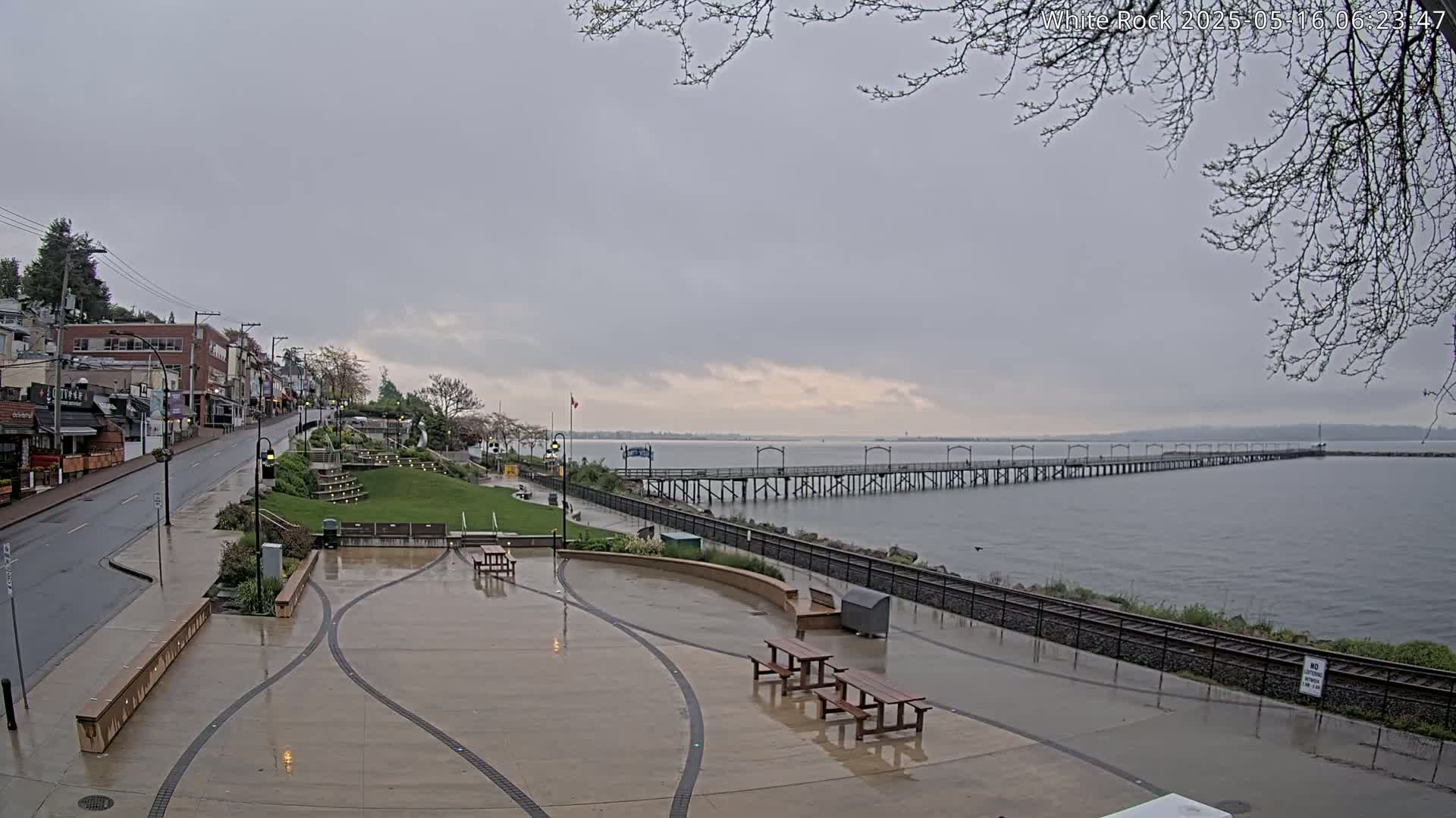A wet, paved waterfront area with picnic tables and benches overlooks a long pier extending into a calm, gray body of water under an overcast sky.