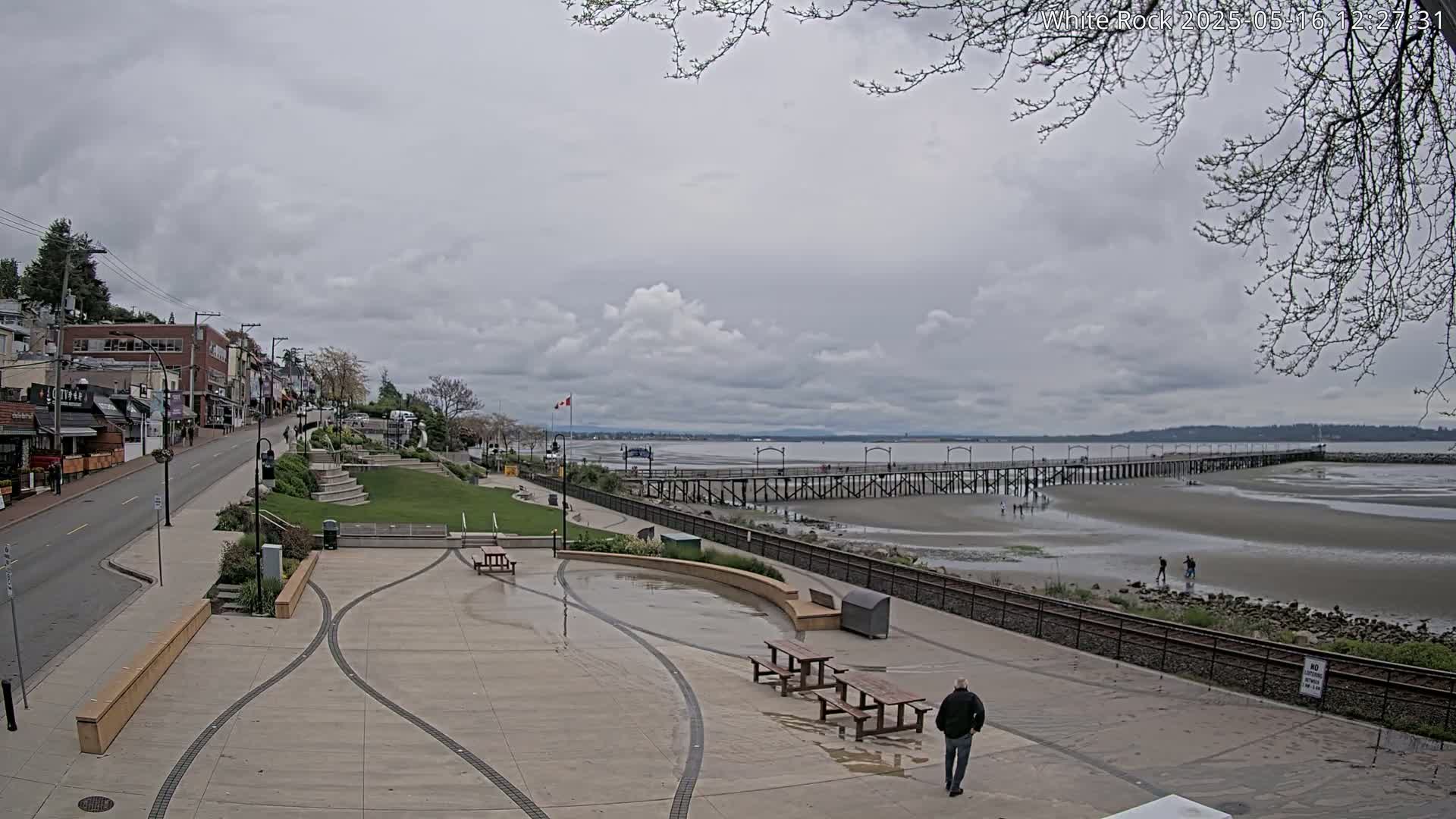 A paved waterfront plaza with picnic tables overlooks a long pier extending into a partially exposed sandy bay under a cloudy sky.