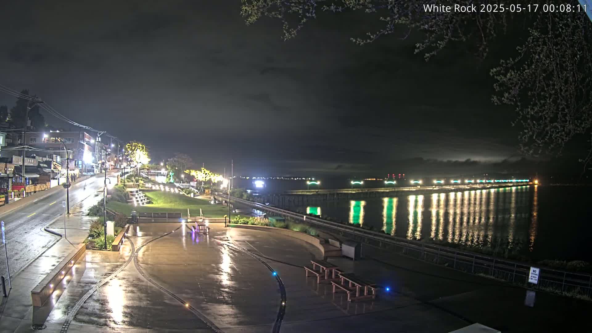A rain-slicked waterfront plaza at night, with illuminated buildings and a pier stretching across a calm, light-reflecting body of water.