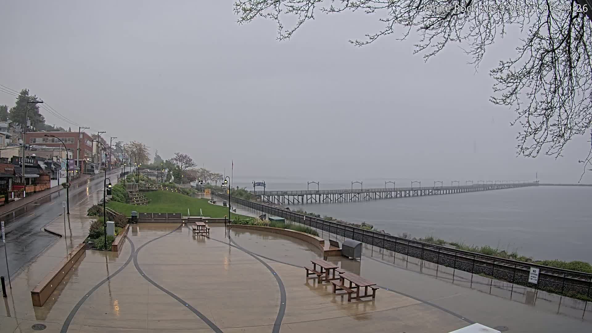 A long pier stretches across a calm body of water under a gray, rainy sky, with a wet town and plaza in the foreground.