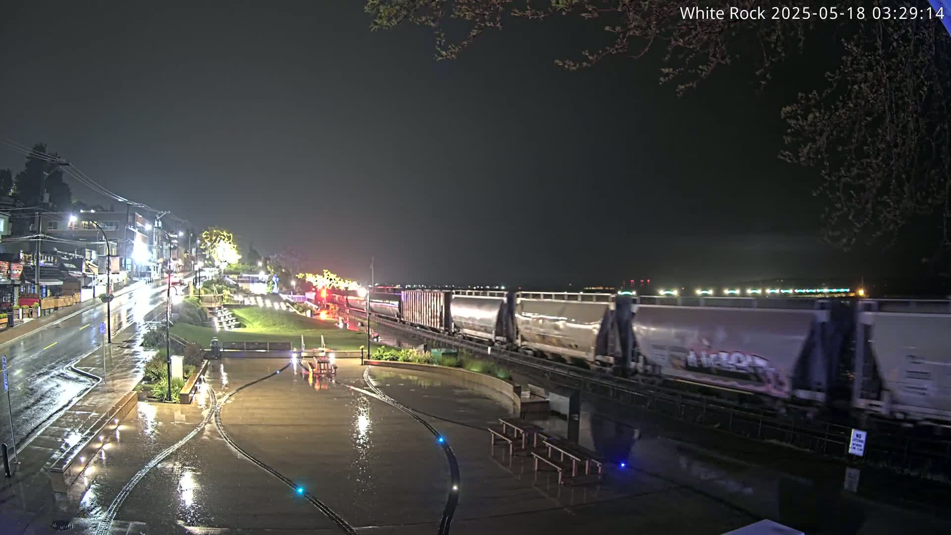 A long freight train passes by a rain-slicked waterfront plaza at night, under a dark sky.