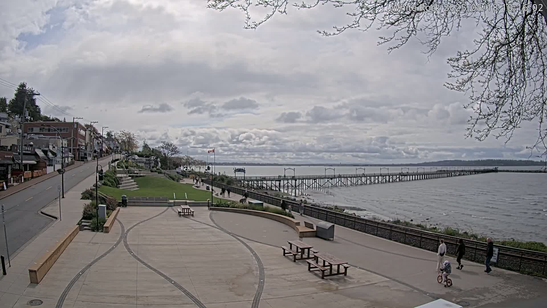 A long pier extends into a calm, cloudy-sky day ocean beside a paved waterfront area with picnic tables and a street lined with buildings.