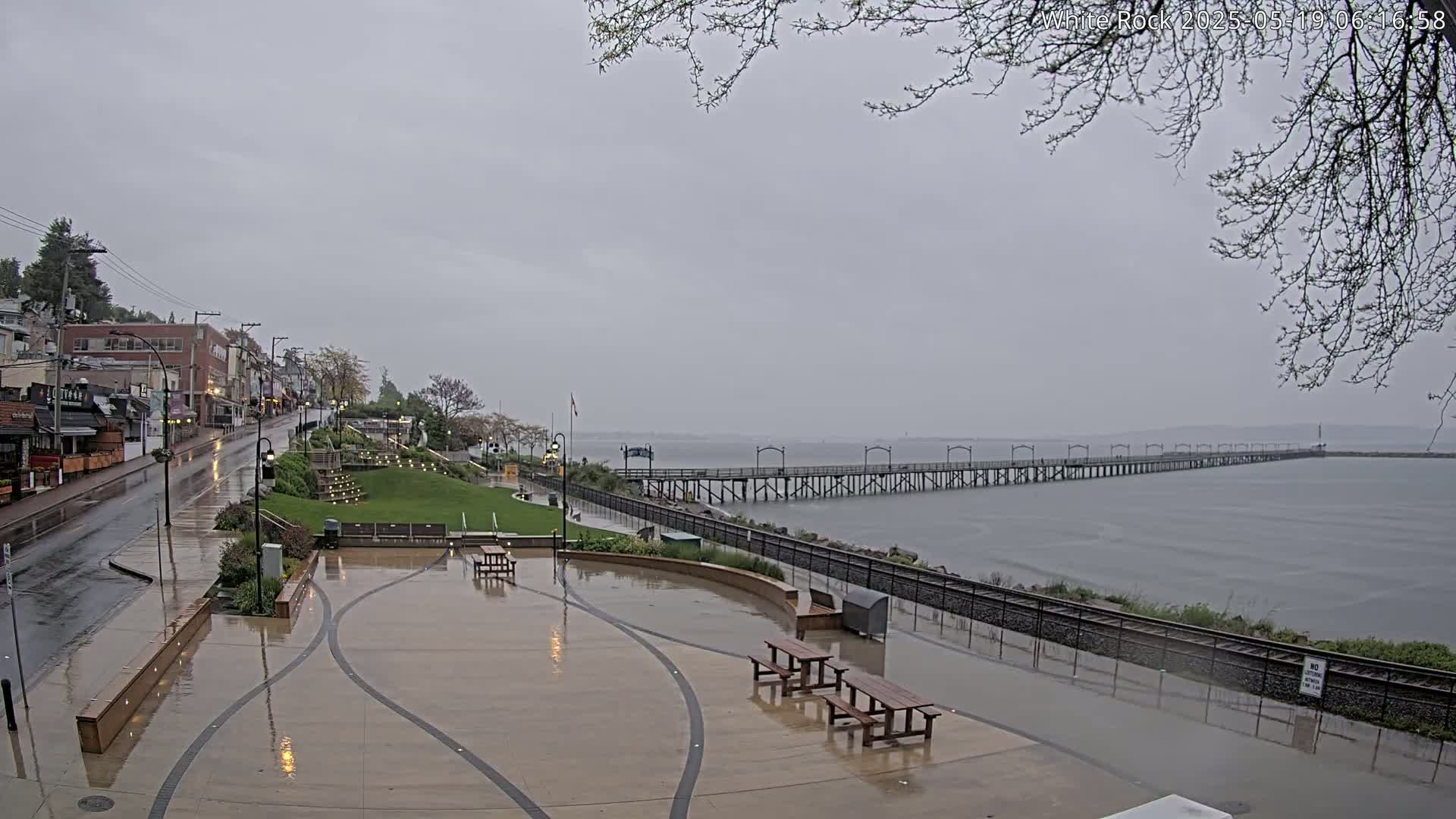 A long pier extends into a calm gray body of water under an overcast sky, overlooking a rain-slicked waterfront plaza and a street lined with buildings.