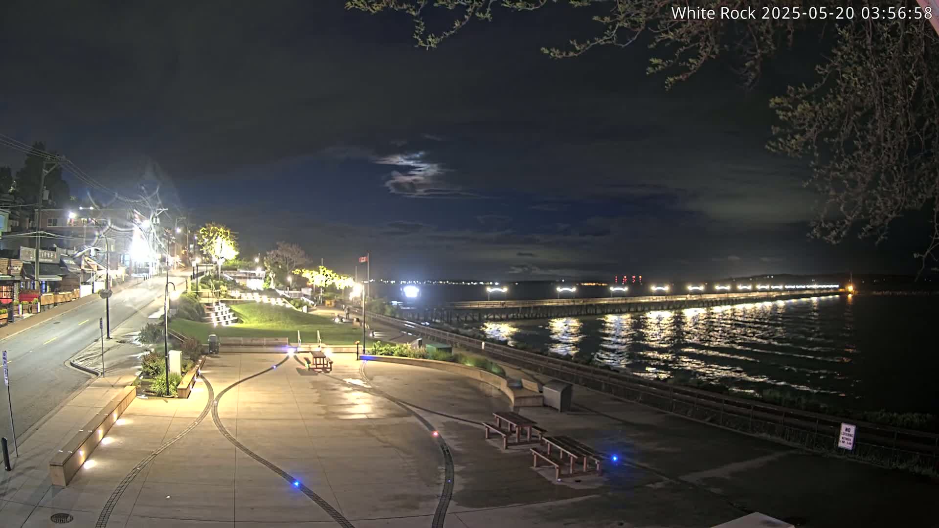 A nighttime view of a waterfront plaza with a long pier extending into a calm body of water under a partly cloudy sky.