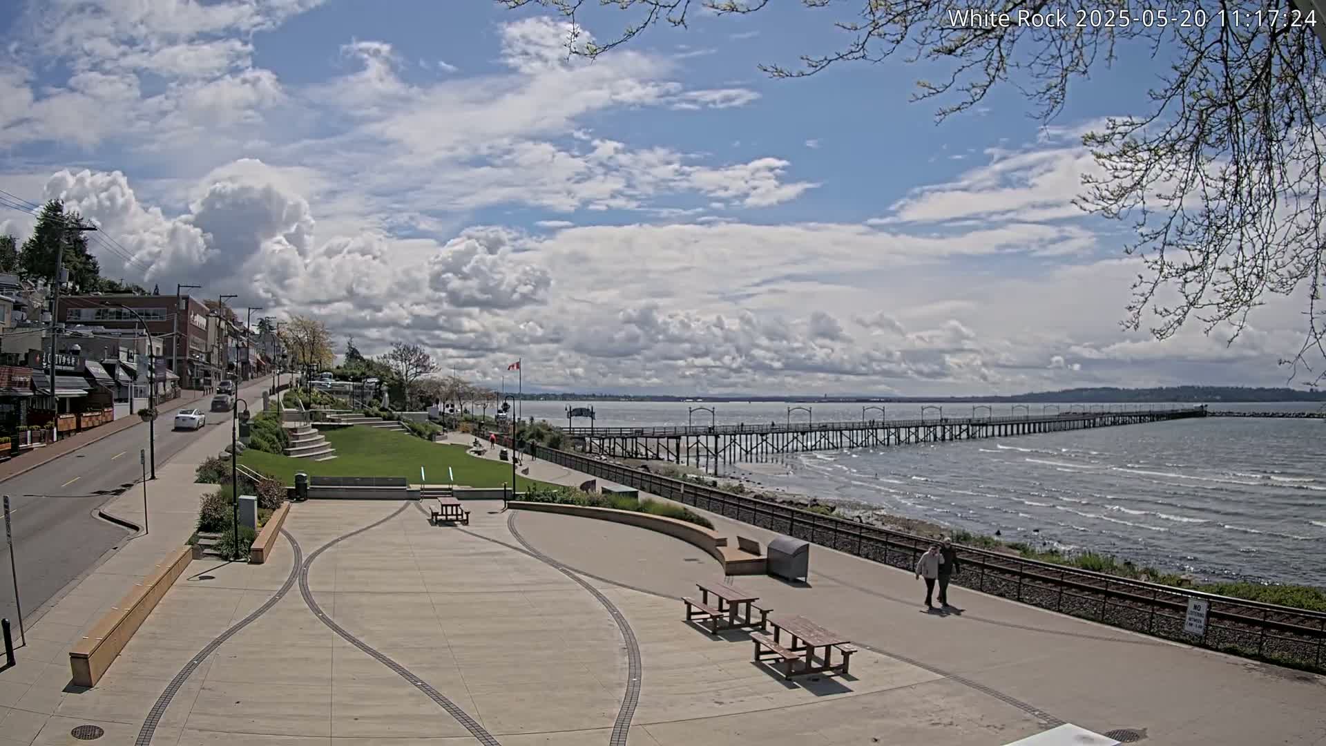 A paved waterfront plaza with picnic tables overlooks a long pier extending into a calm, wavy ocean under a partly cloudy sky.