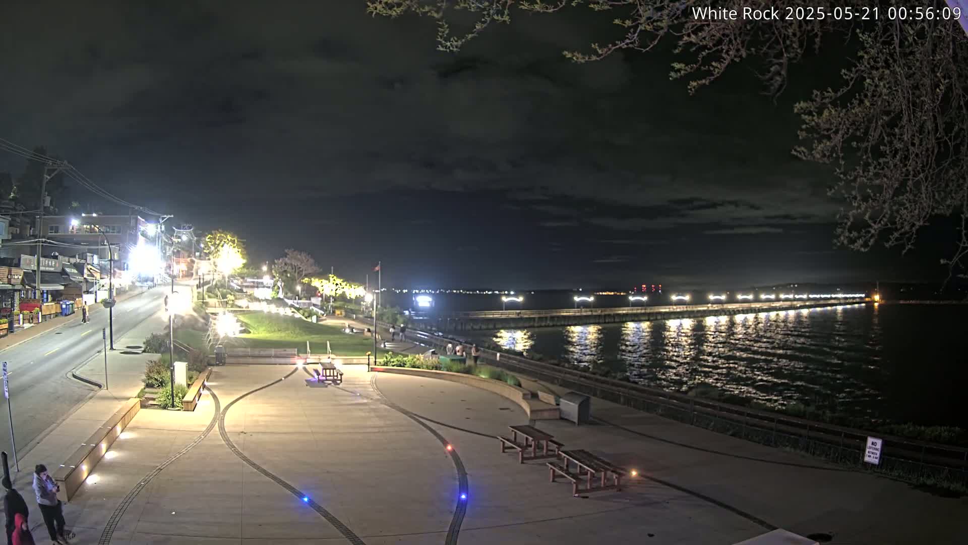 A nighttime view of a waterfront plaza with a pier extending into a calm, dark body of water under a partly cloudy sky.