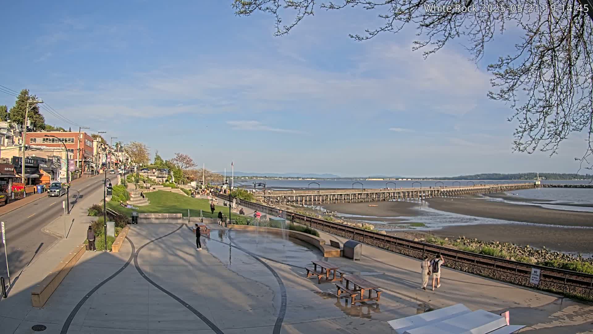 A waterfront town scene on a partly sunny day shows a long pier extending into a calm bay, with a paved plaza and grassy area in the foreground populated by a few people.