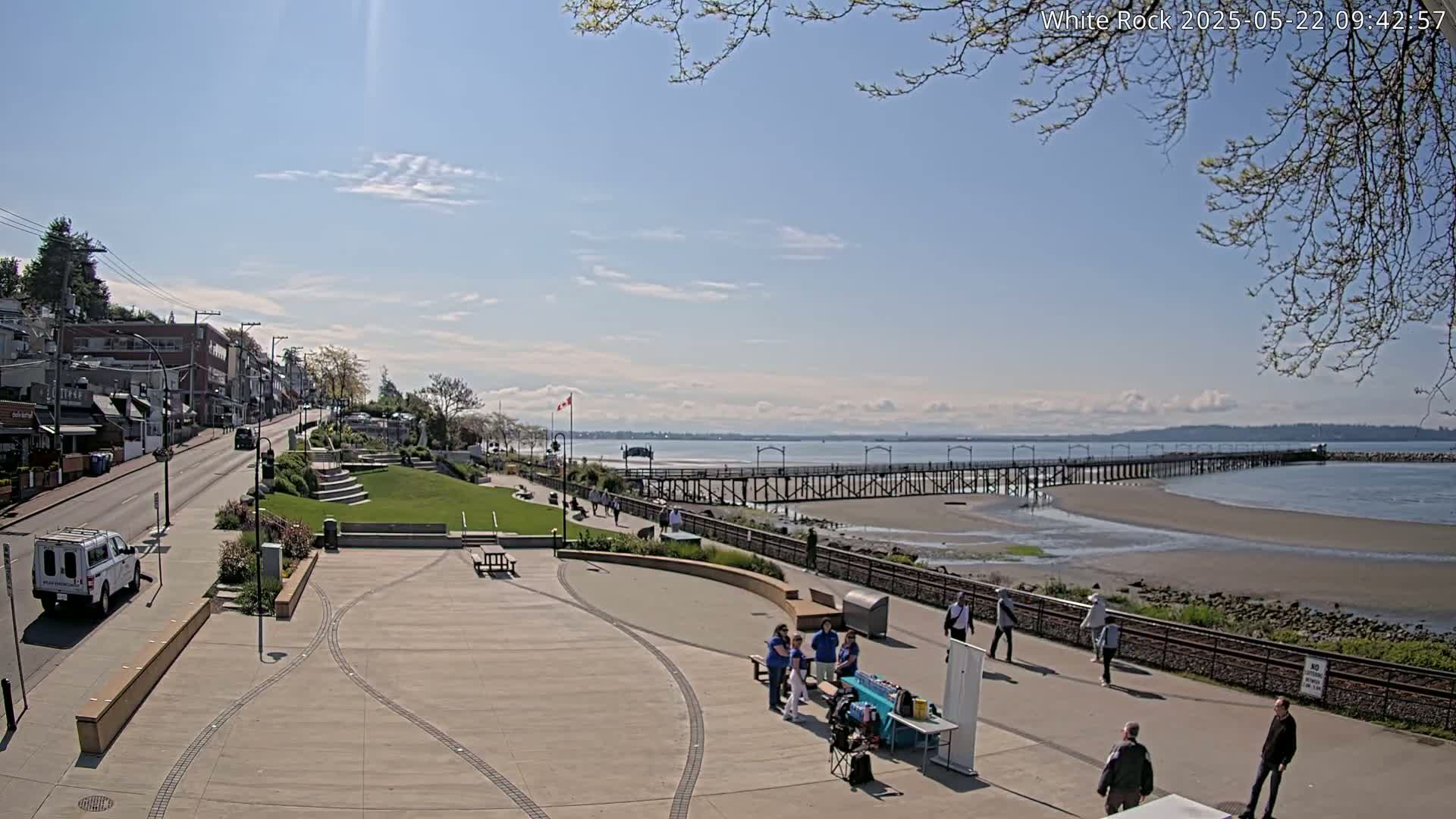A sunny day reveals a seaside town with a long pier extending into calm water, people strolling along a paved walkway, and buildings lining a street.
