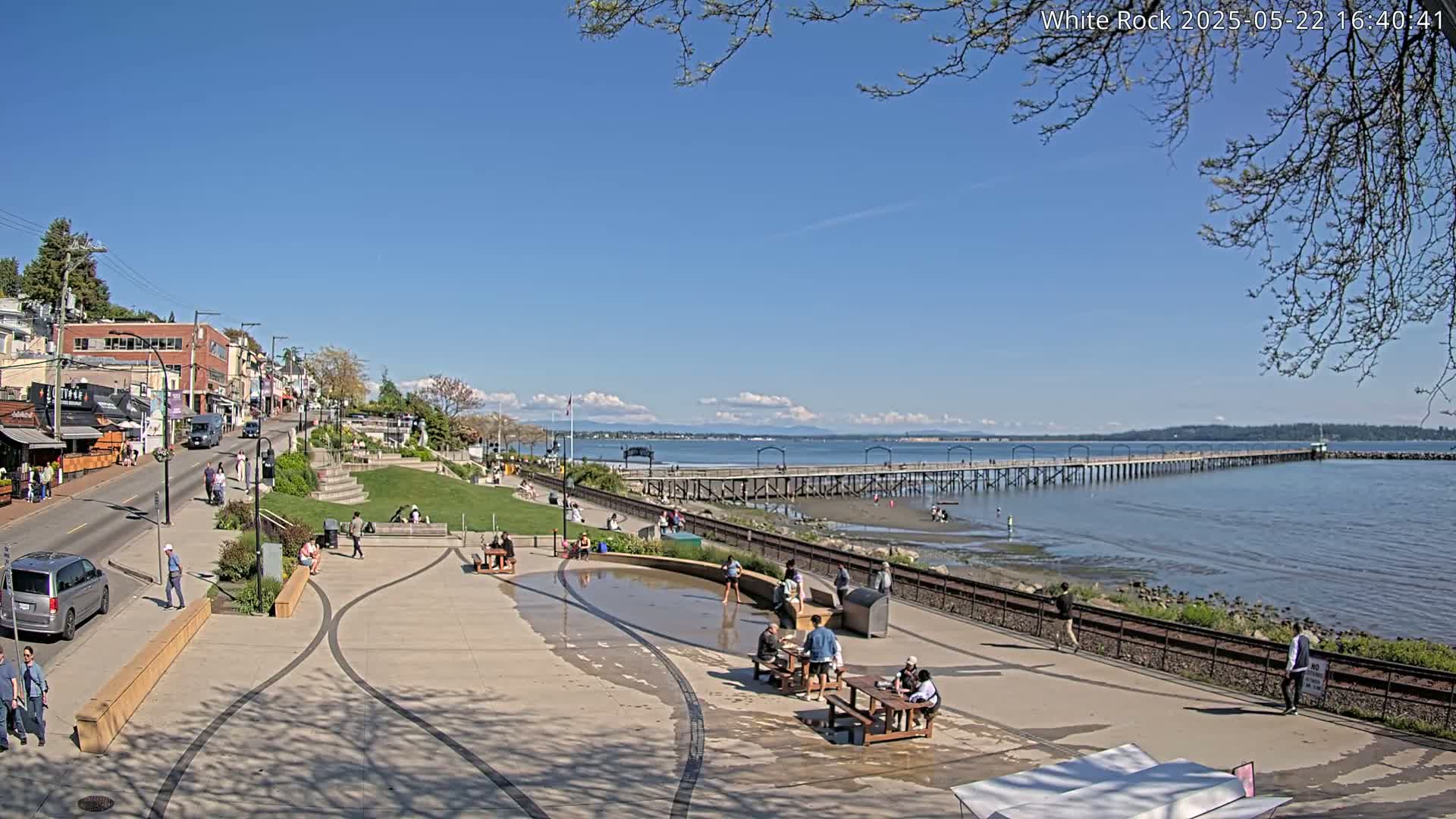 A sunny day reveals a waterfront town with a long pier extending into the calm sea, people enjoying the park and promenade.