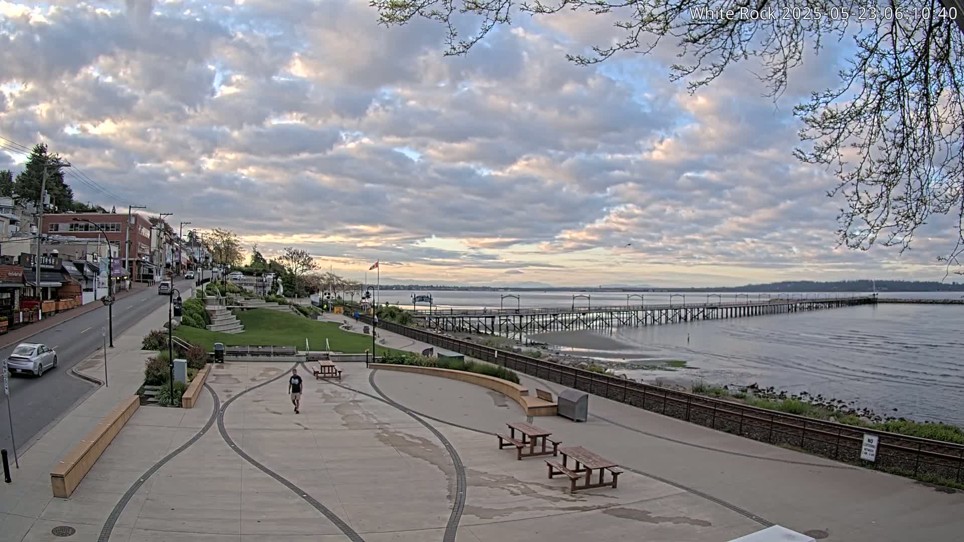 A long pier extends into a calm body of water under a partly cloudy sky, with a paved waterfront area featuring picnic tables and a person walking, and buildings and a road visible in the background.