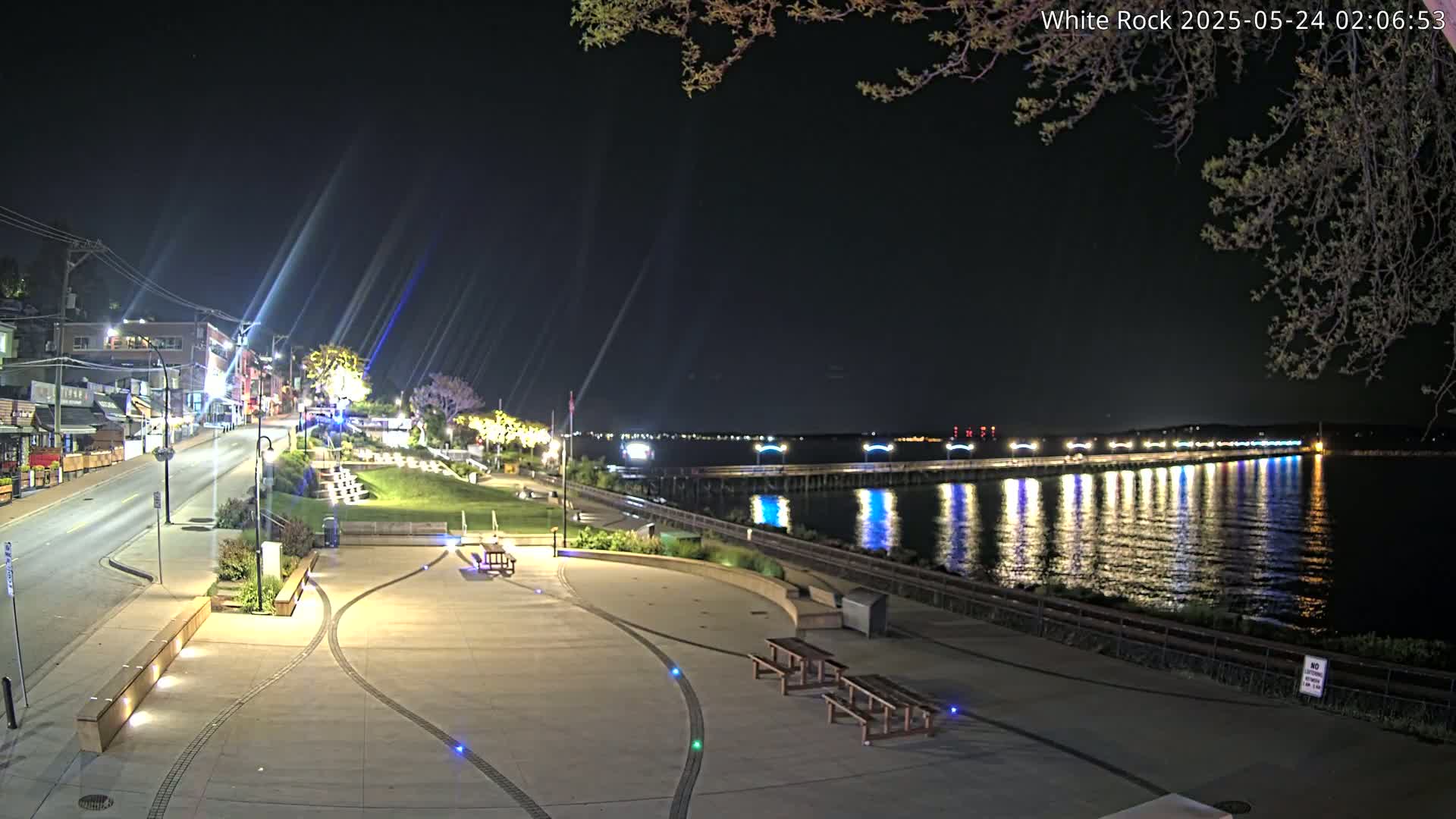 A nighttime view of a waterfront plaza with benches and a pier extending into a calm, illuminated body of water under a clear, dark sky.