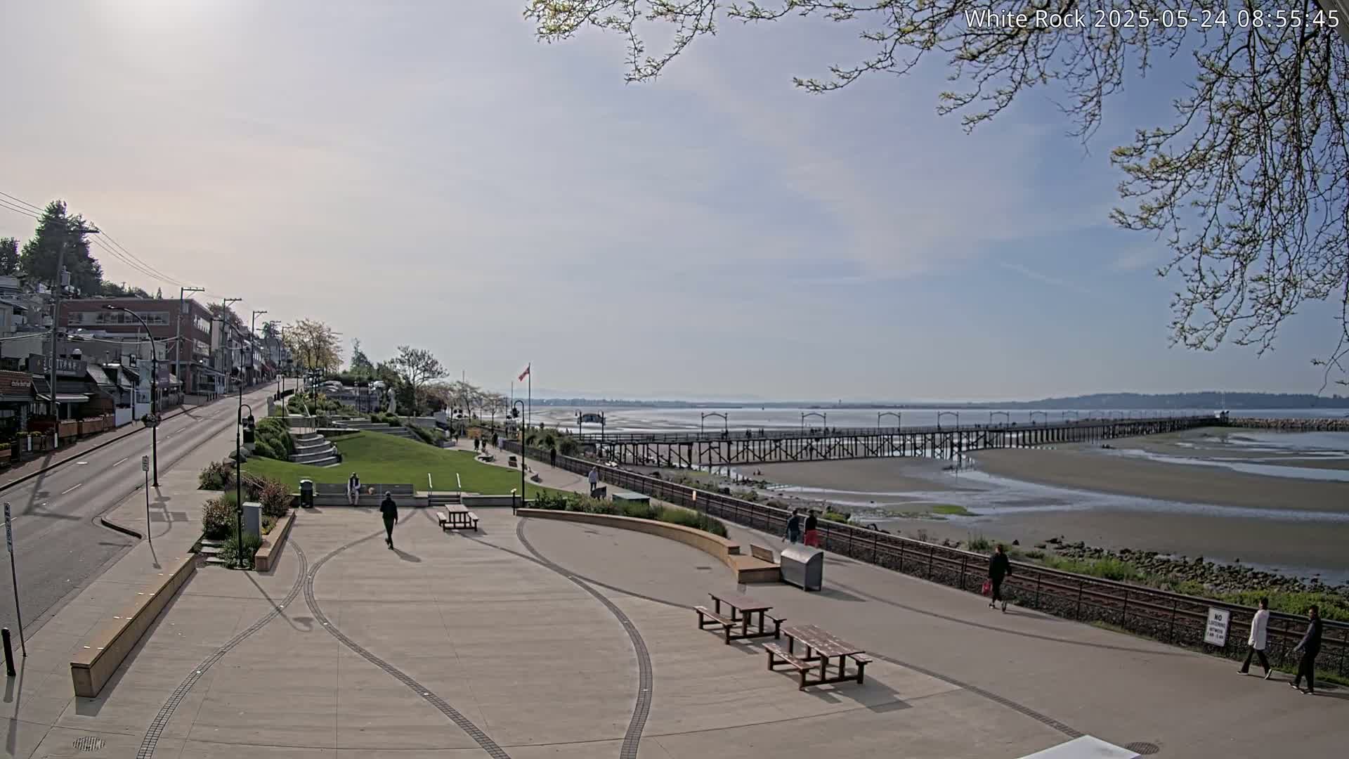 A paved waterfront area with picnic tables and a long pier extending into a calm, partially exposed bay under a mostly sunny sky.