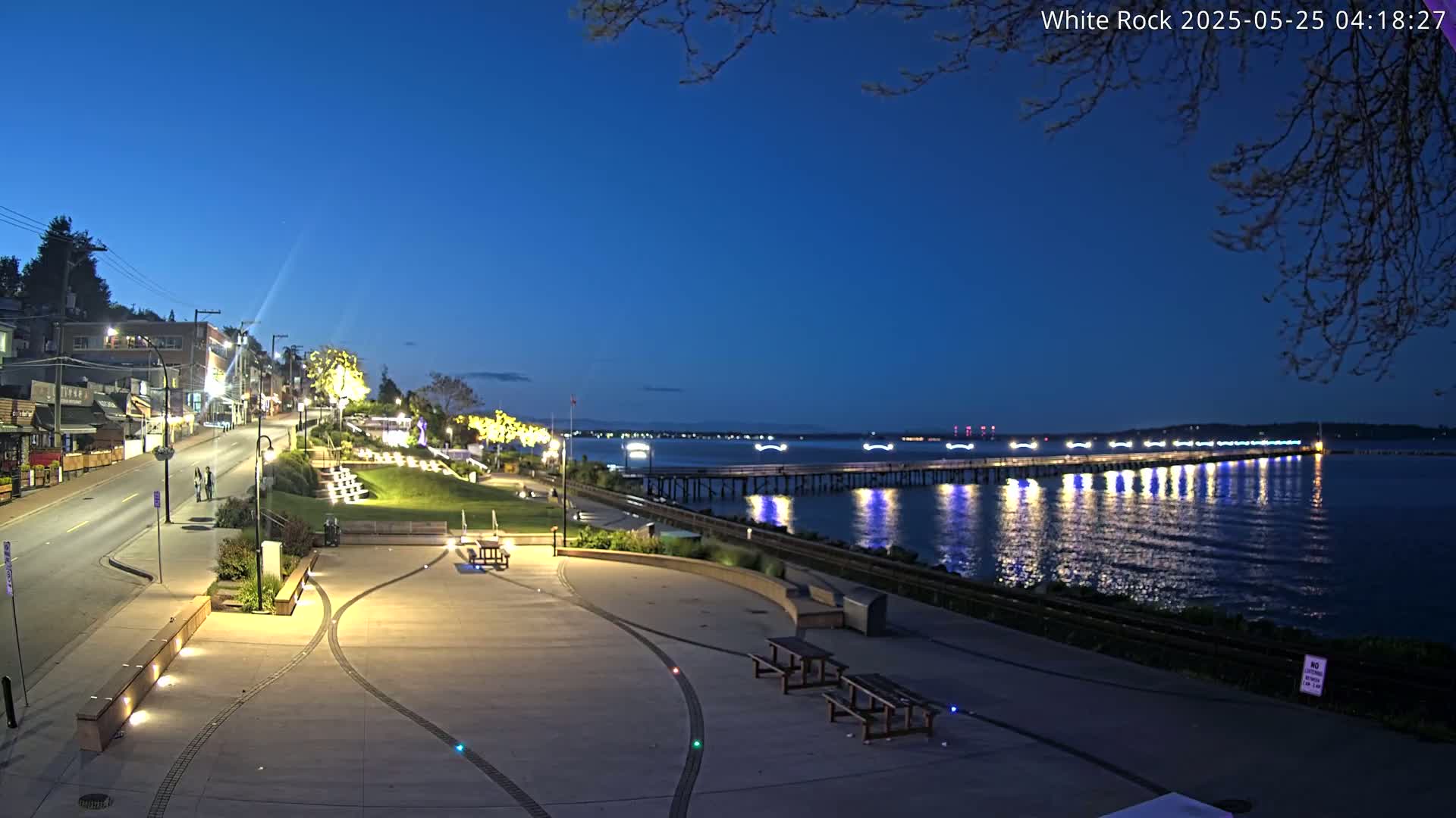 A waterfront plaza at twilight is illuminated, showcasing a long pier extending into the calm, dark blue water under a clear sky.