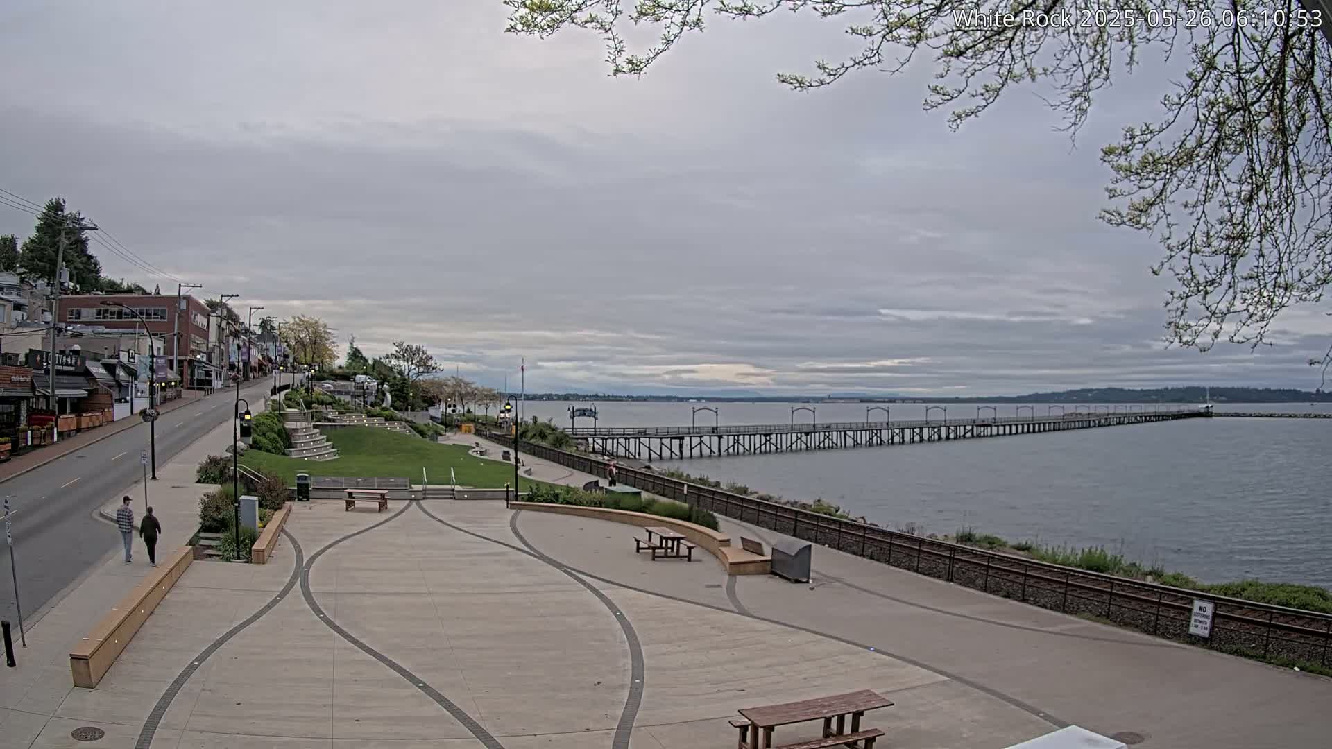 A long pier extends into a calm bay under an overcast sky, with a paved waterfront area and a street lined with buildings visible in the foreground.