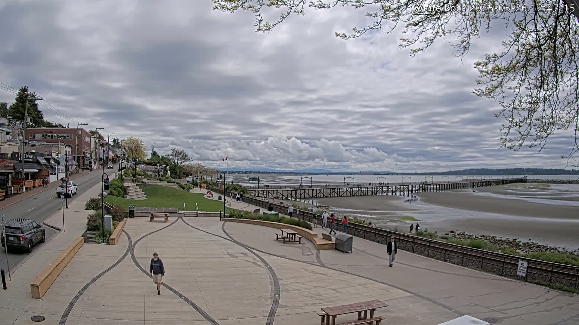 A paved waterfront plaza with a long pier extending into a tidal flat under a cloudy sky.