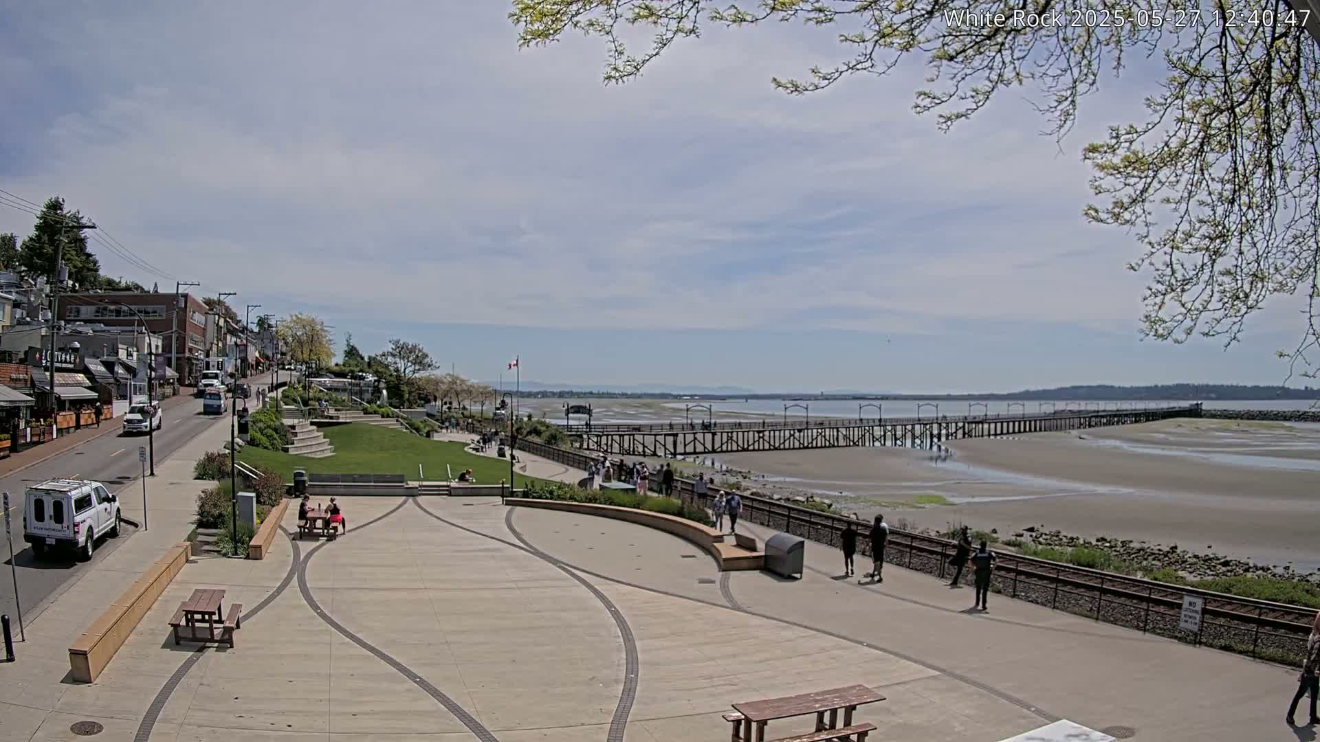 A paved waterfront plaza with picnic tables and a long pier extending into a sandy, low-tide beach under a partly cloudy sky.