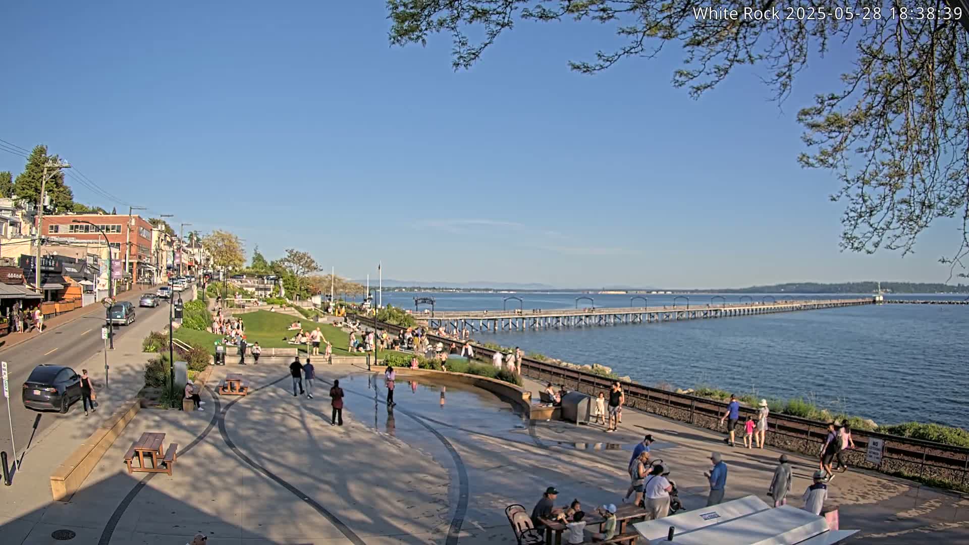 A sunny waterfront scene shows a long pier extending into calm blue water, with many people enjoying a paved plaza and grassy area near shops and a road.