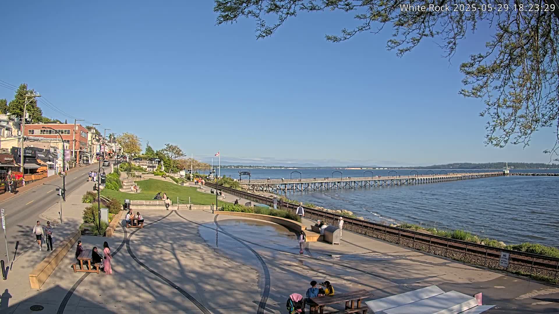 A seaside town with a long pier extending into calm, blue water under a clear, sunny sky, features a paved waterfront area with people relaxing and enjoying the view.