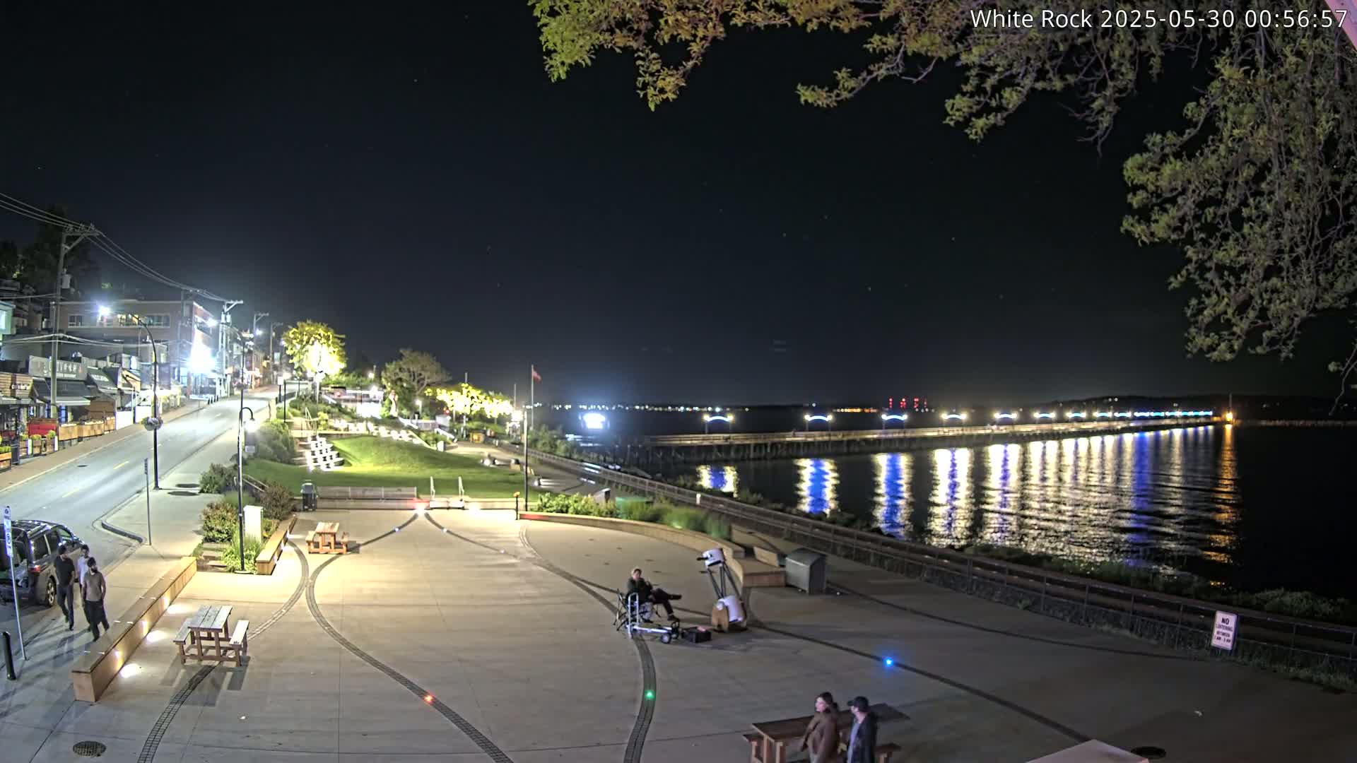 A nighttime view of a waterfront plaza with a long pier extending into a calm, illuminated body of water, under a clear, starry sky.