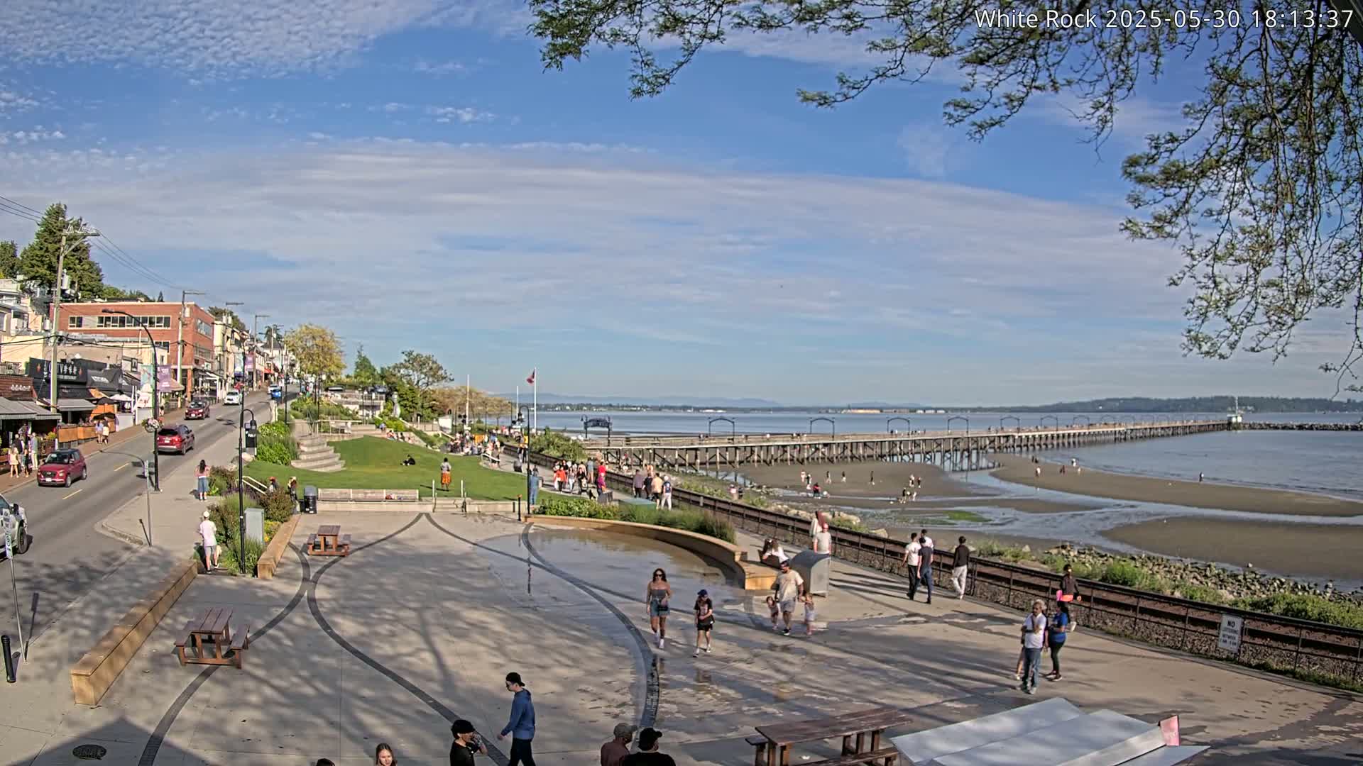 A seaside town square with a long pier extending into the calm water under a partly cloudy sky, filled with people strolling and enjoying the sunny weather.