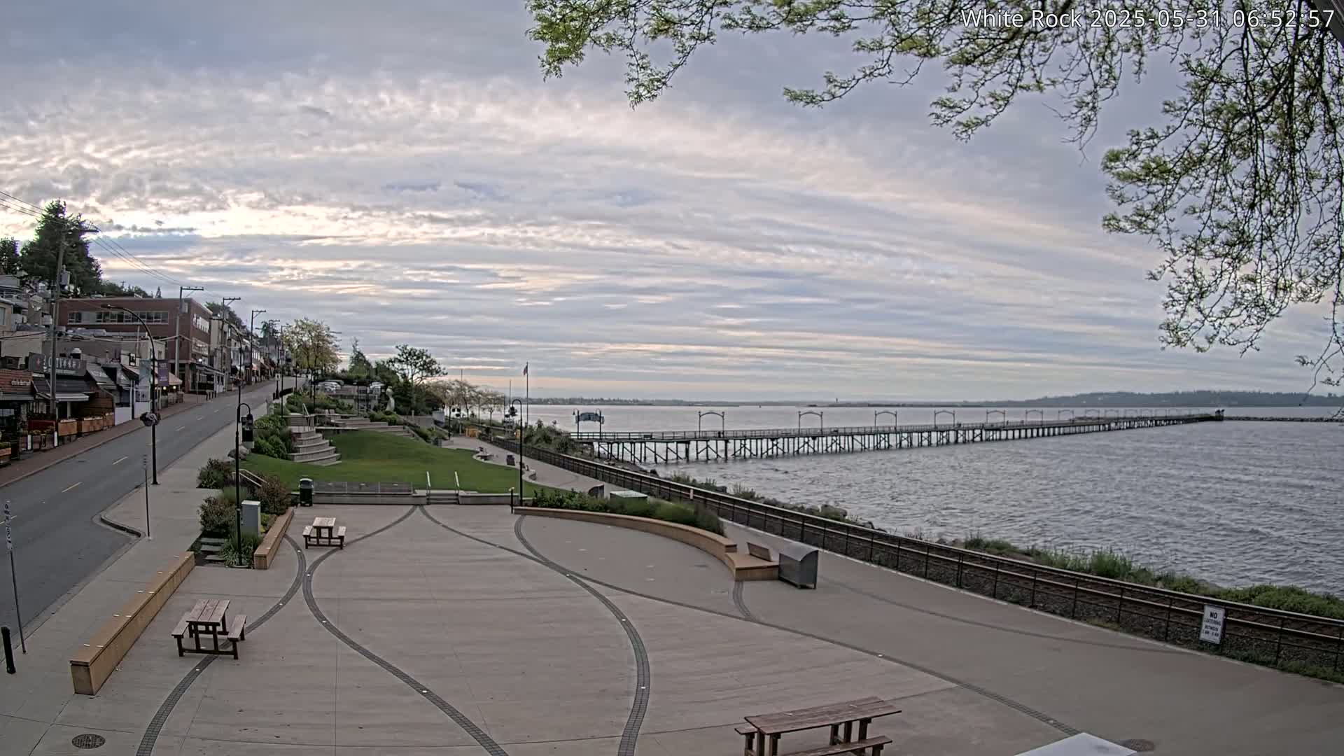 A paved waterfront plaza with picnic tables overlooks a long pier extending into a calm, overcast body of water, with a street and buildings visible in the background.