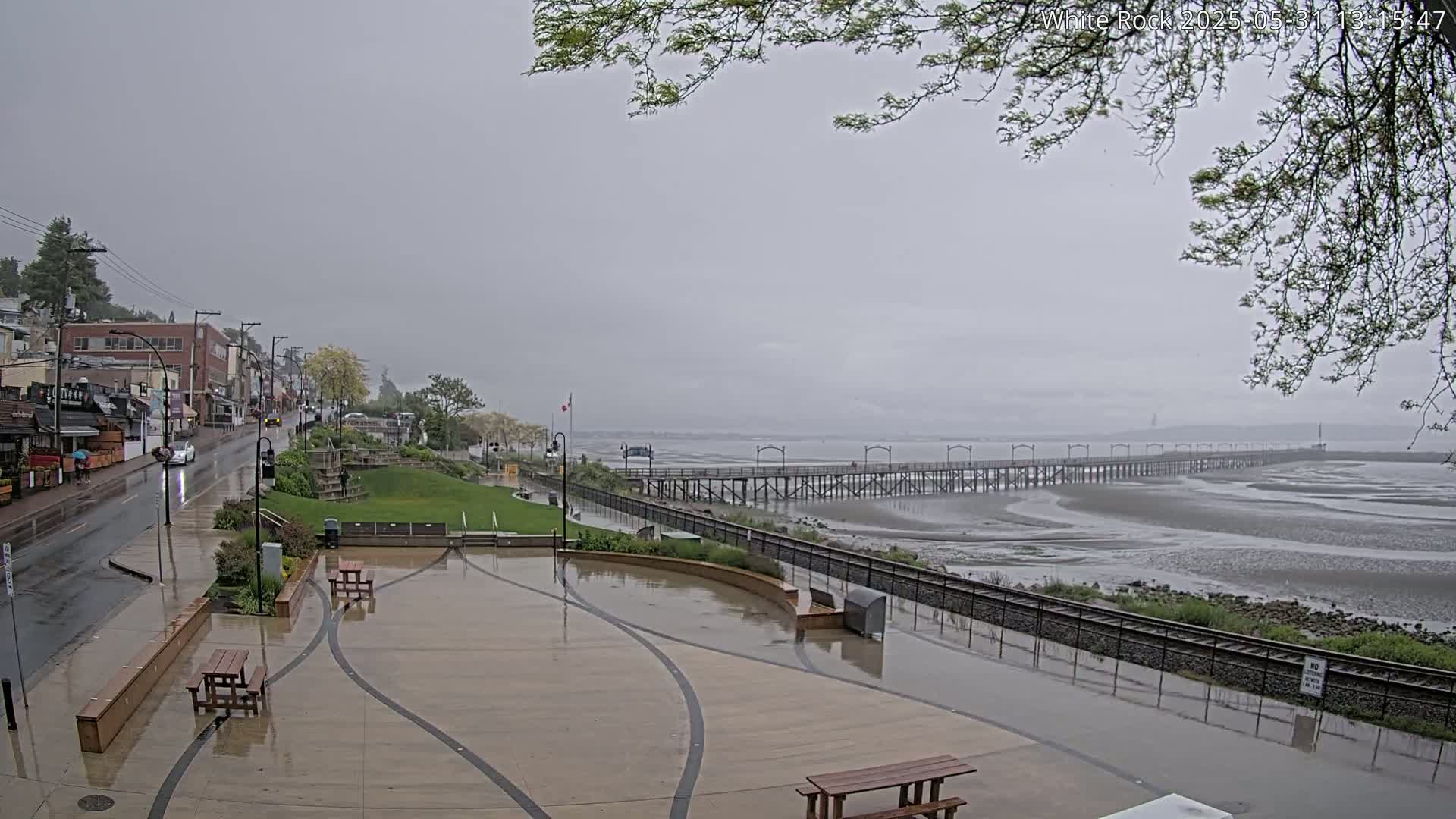 A wet, paved plaza with picnic tables overlooks a long pier extending into a calm, gray body of water under an overcast sky.