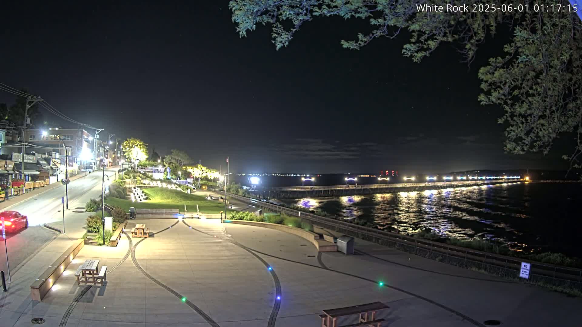 A nighttime view of a waterfront plaza with a road and buildings behind it, overlooking a calm body of water with a pier in the distance under a mostly clear sky.