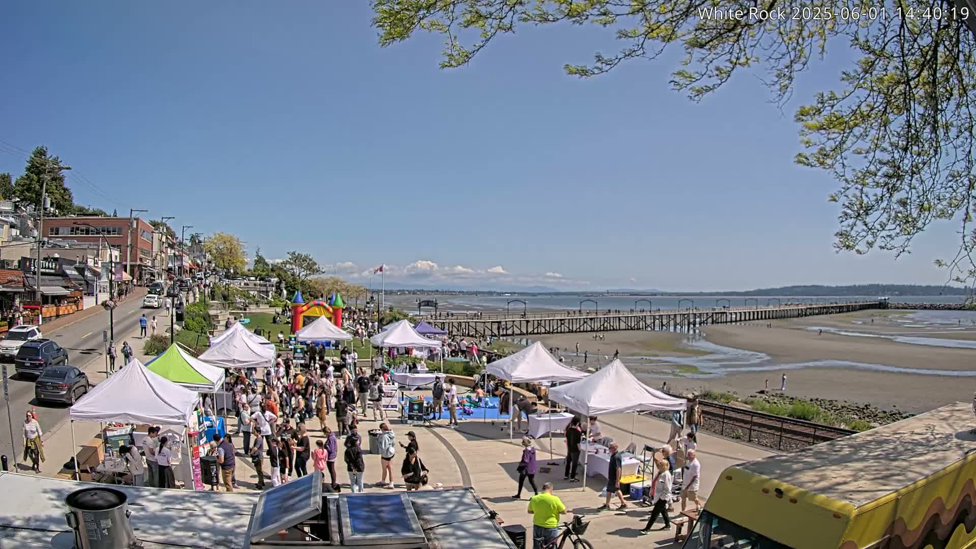 A sunny day reveals a seaside town with a crowd gathered around market stalls and a bouncy castle near a long pier, extending into a wide, exposed bay.