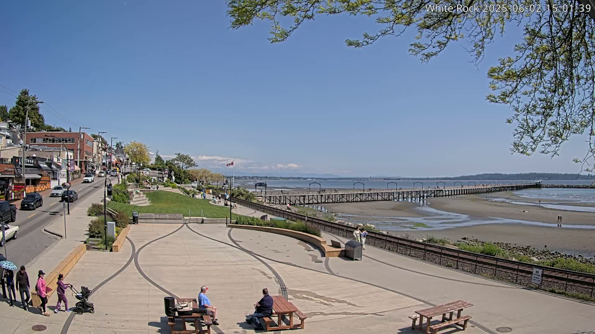 A sunny day reveals a seaside town with a long pier extending into the calm ocean, a paved plaza with people relaxing, and a street lined with buildings.