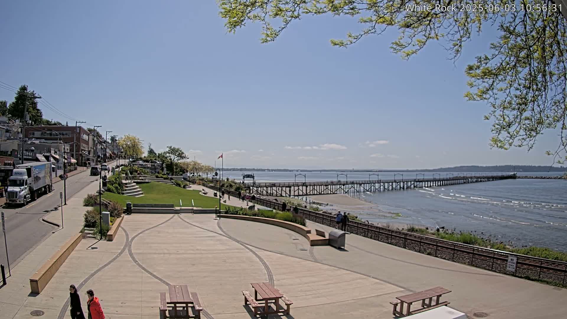 A paved waterfront plaza with picnic tables overlooks a long pier extending into a calm, sunny ocean.