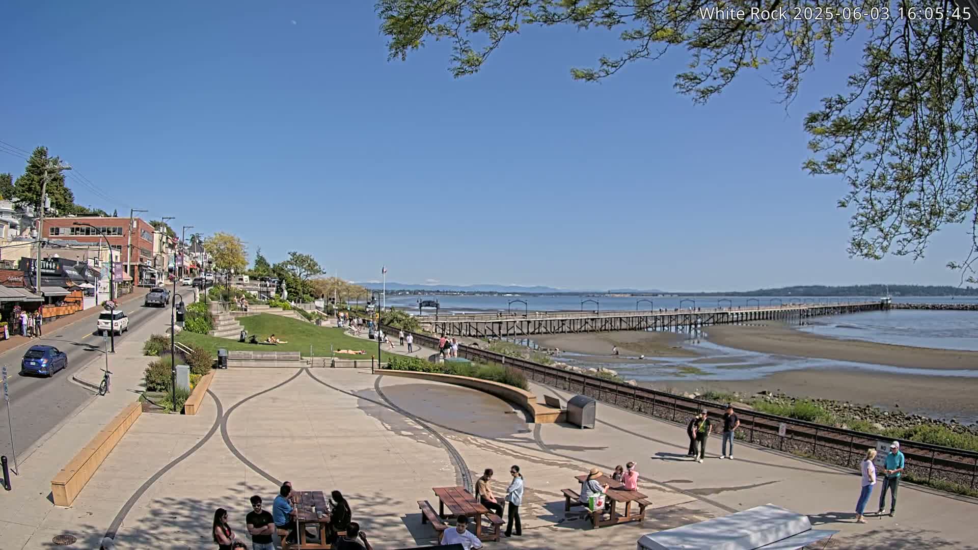 A seaside town square with picnic tables and people relaxing, a long pier extending into a calm bay under a clear blue sky.