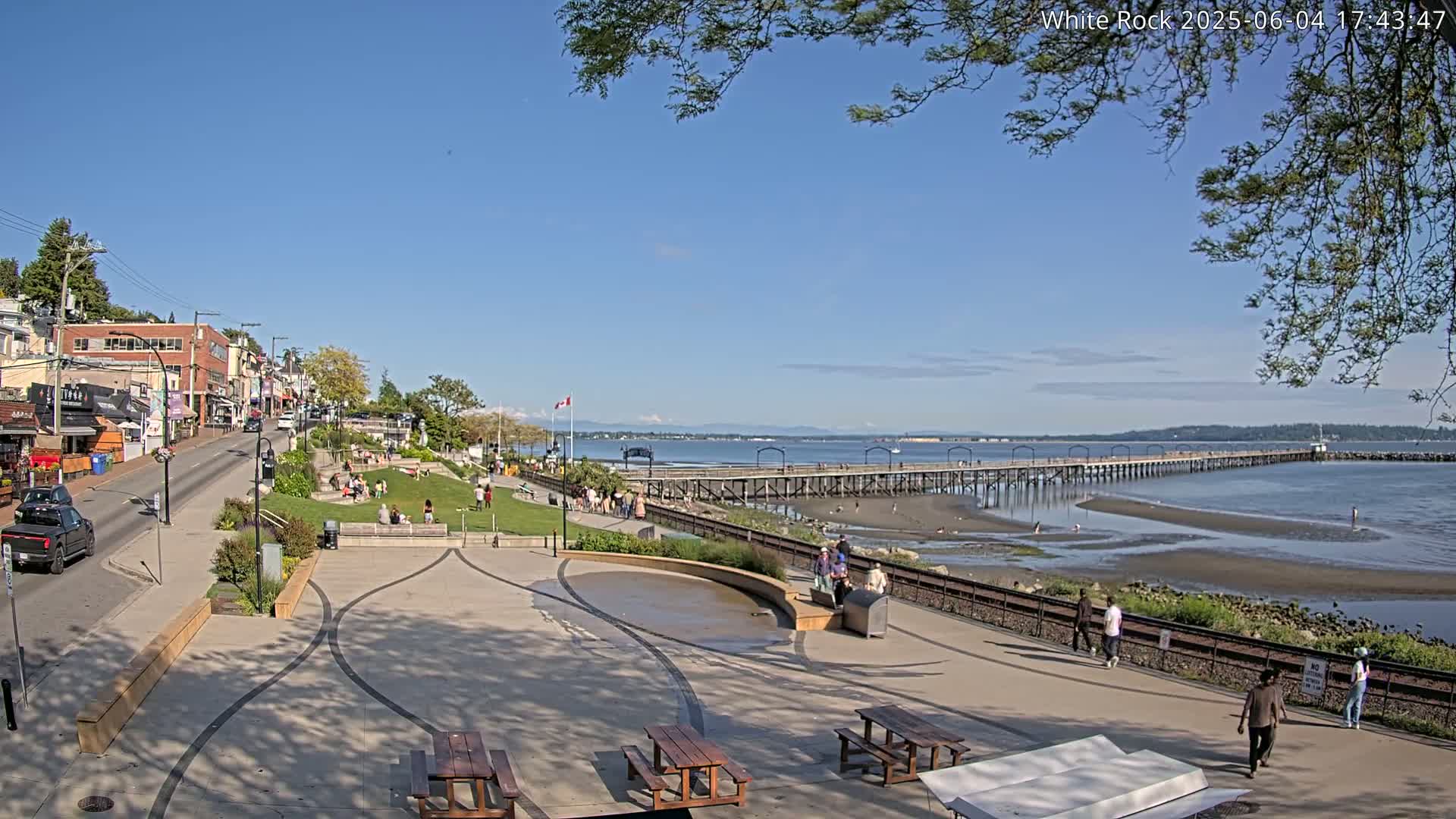 A sunny waterfront scene shows a paved plaza with picnic tables, a grassy area with people relaxing, a long pier extending into calm water, and a street lined with buildings in the background.