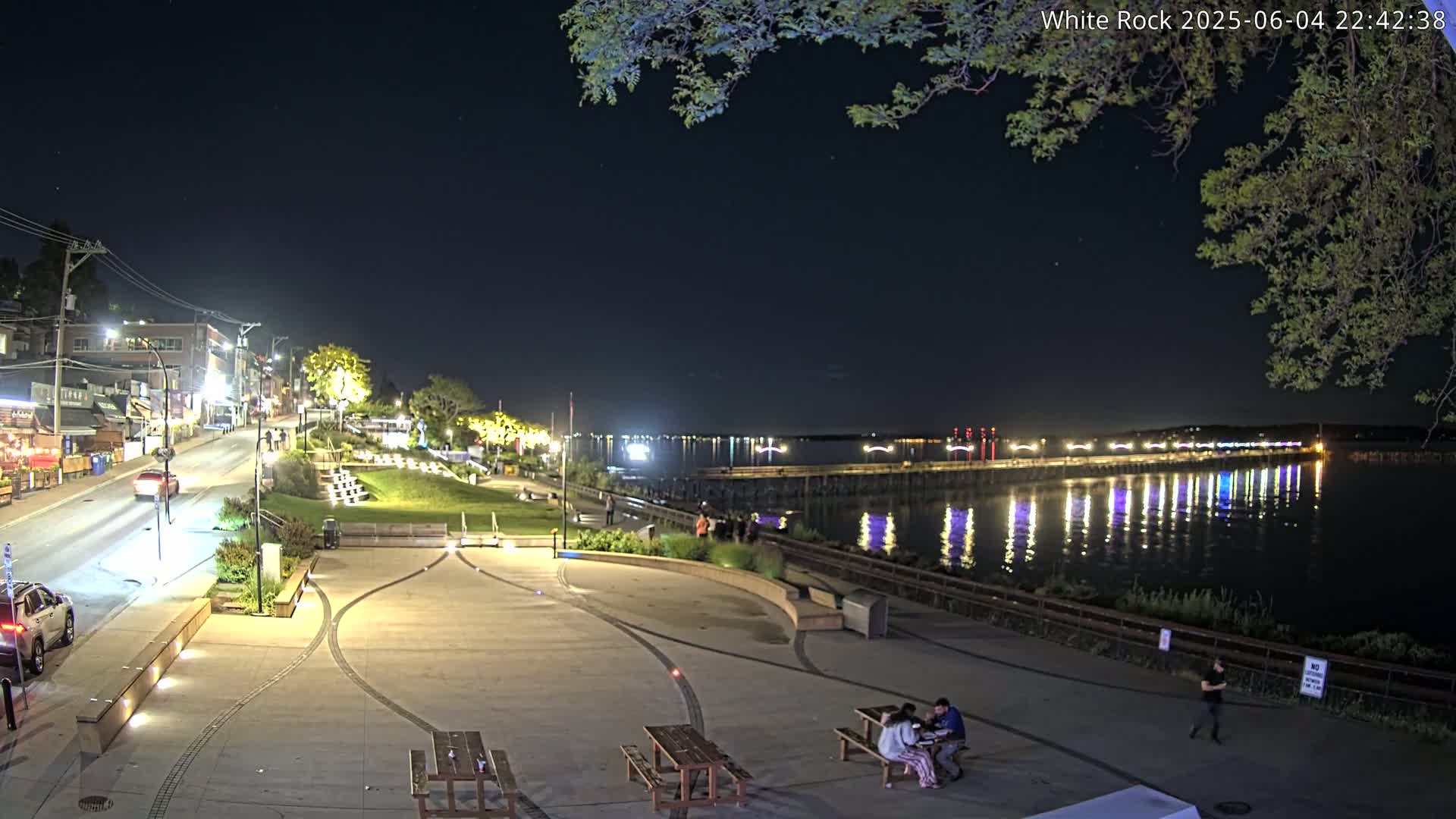 A nighttime view of a waterfront plaza with people sitting at picnic tables, a street with cars, and a long pier extending into a calm body of water reflecting lights.