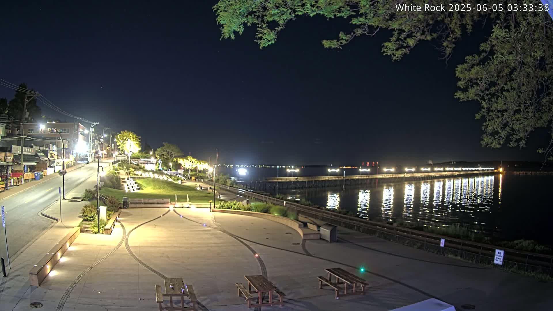 A nighttime view of a waterfront plaza with picnic tables, a street, and buildings, illuminated by lights, overlooking a calm body of water with a pier reflecting the lights.